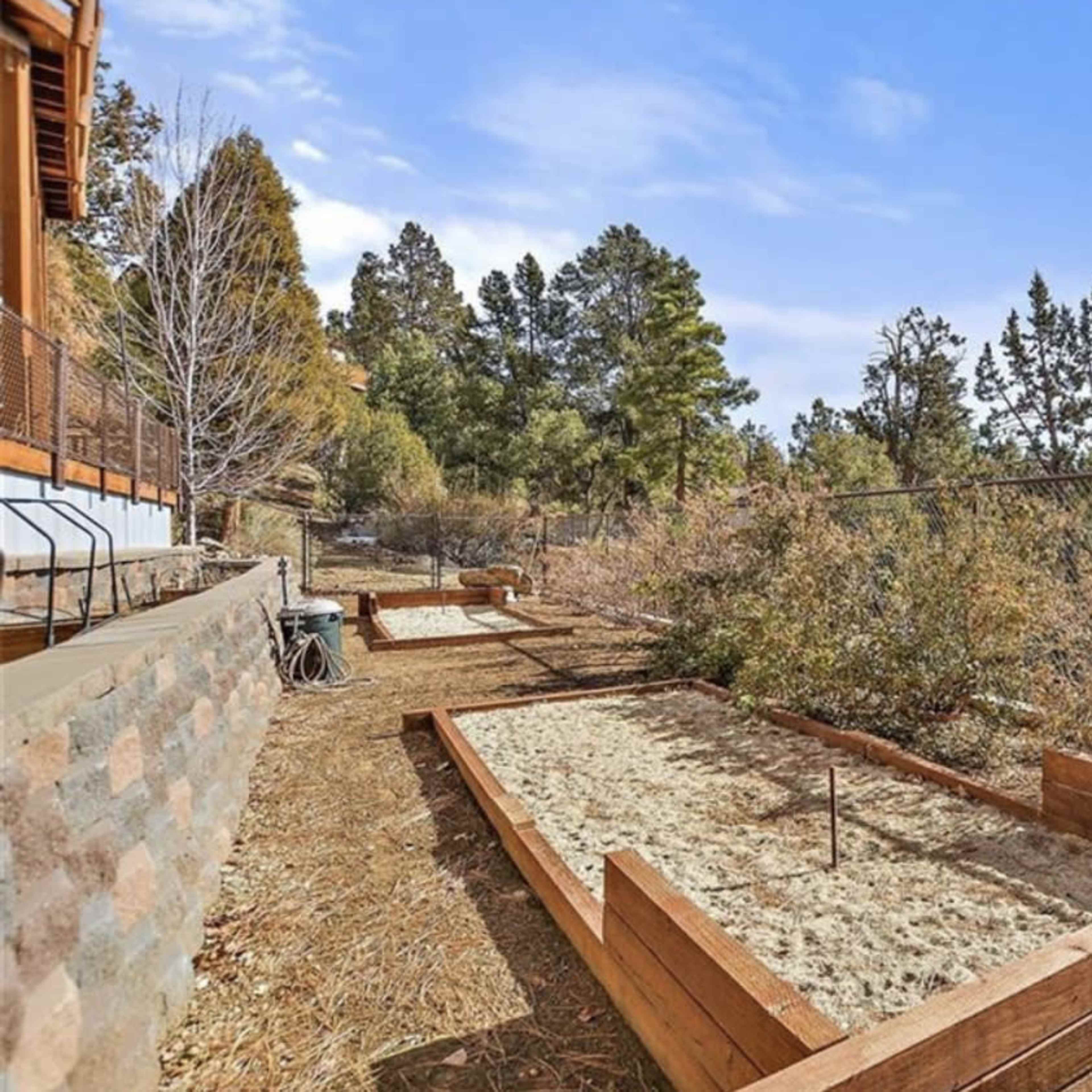 A garden area with wooden raised beds set on a dirt path, surrounded by trees and fencing.