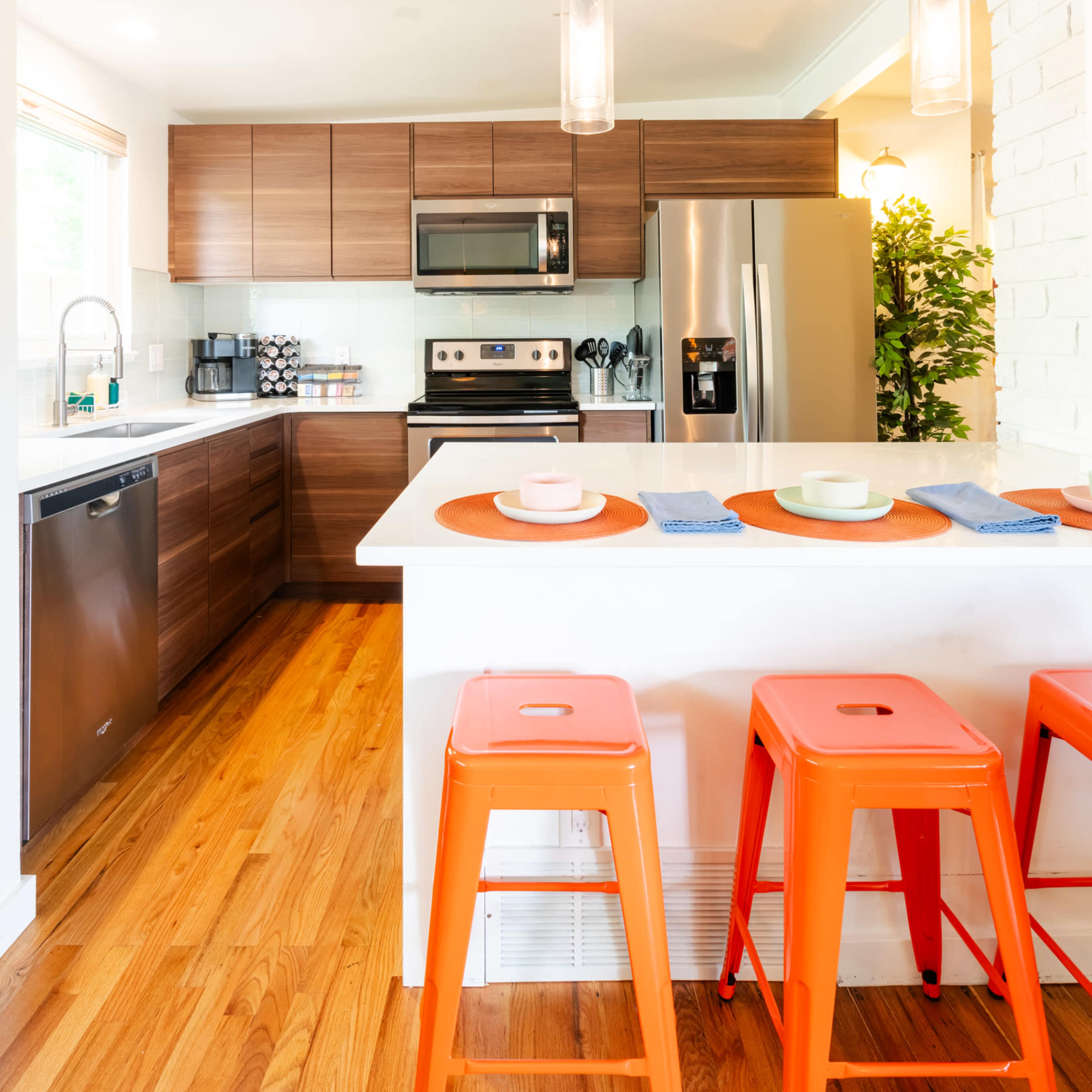 A modern kitchen features wooden cabinets, stainless steel appliances, and orange bar stools at a white countertop.
