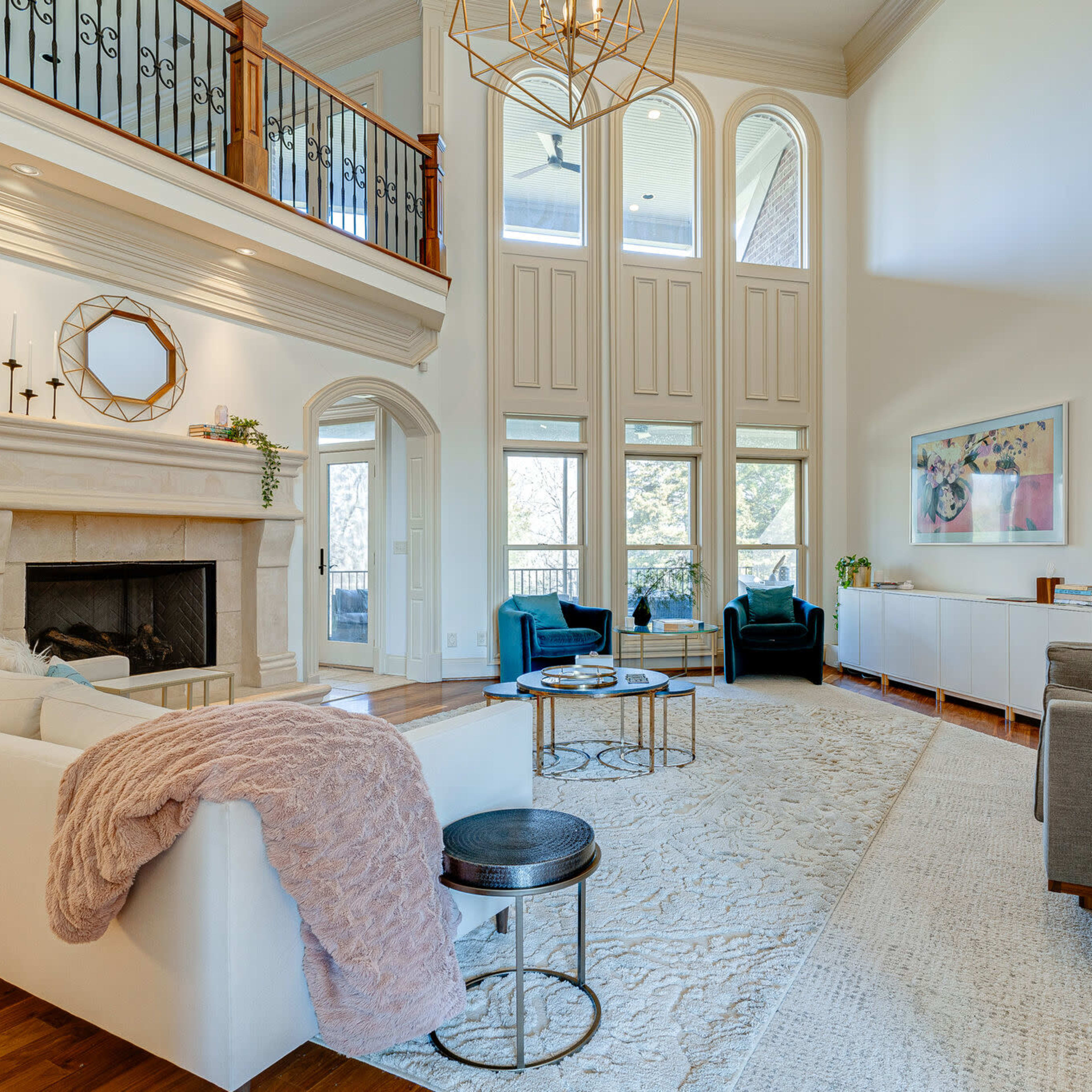 A spacious living room with high ceilings, featuring a stone fireplace, large windows, and modern furniture arranged around a central area rug.