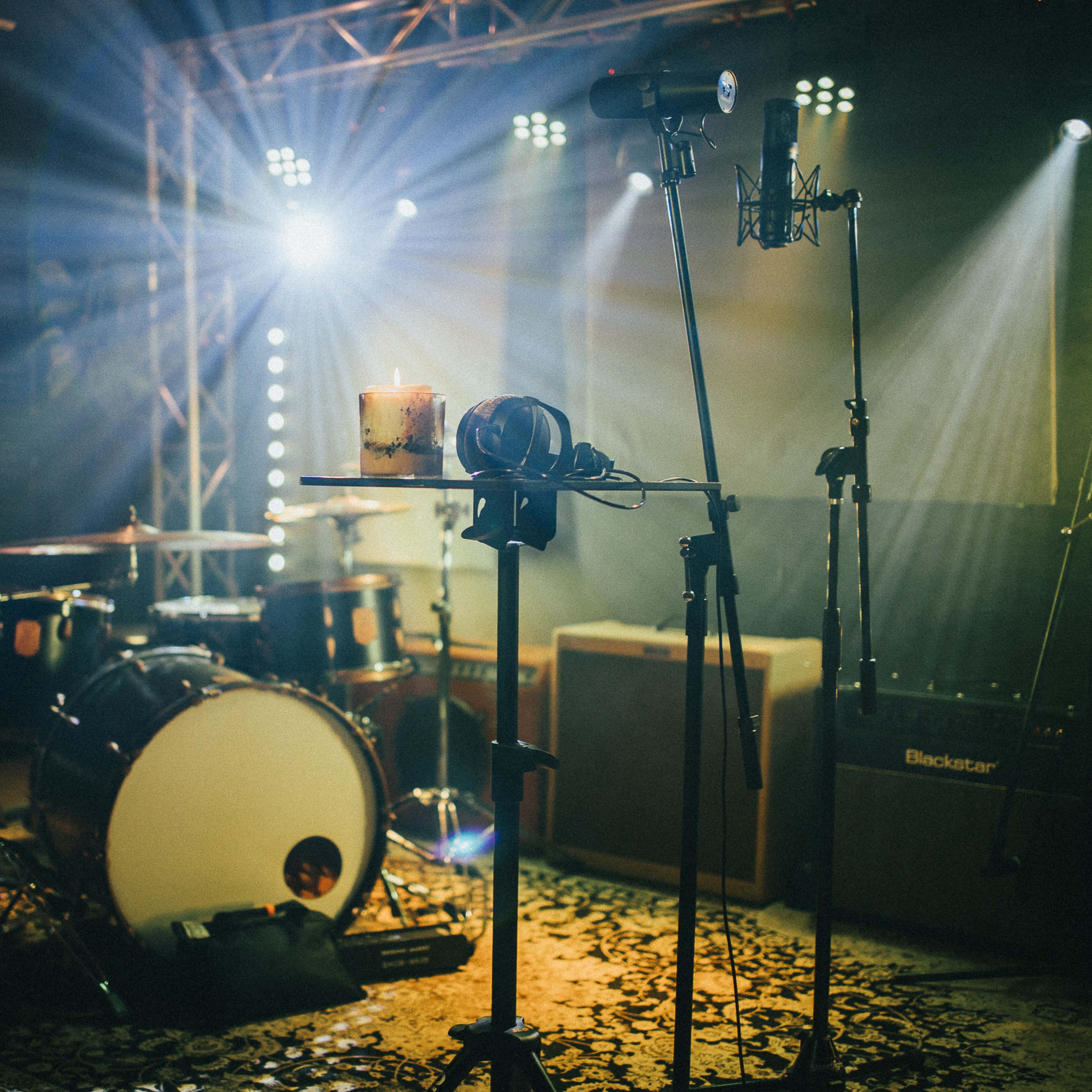 A music studio setup with a drum kit, microphones, a drink on a stand, and various instruments illuminated by stage lights.