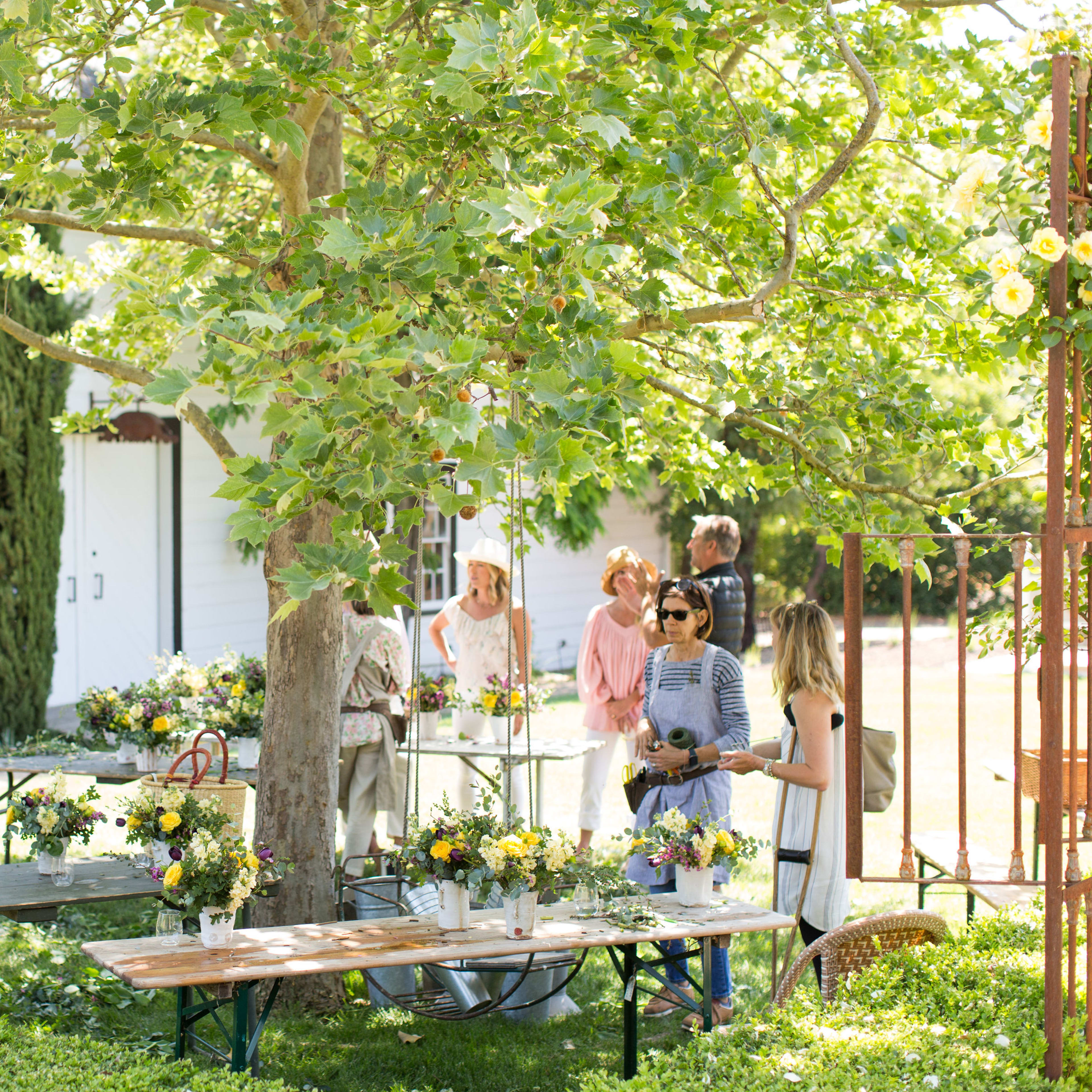 A group of people arrange flower bouquets on tables under a large tree in a garden setting.