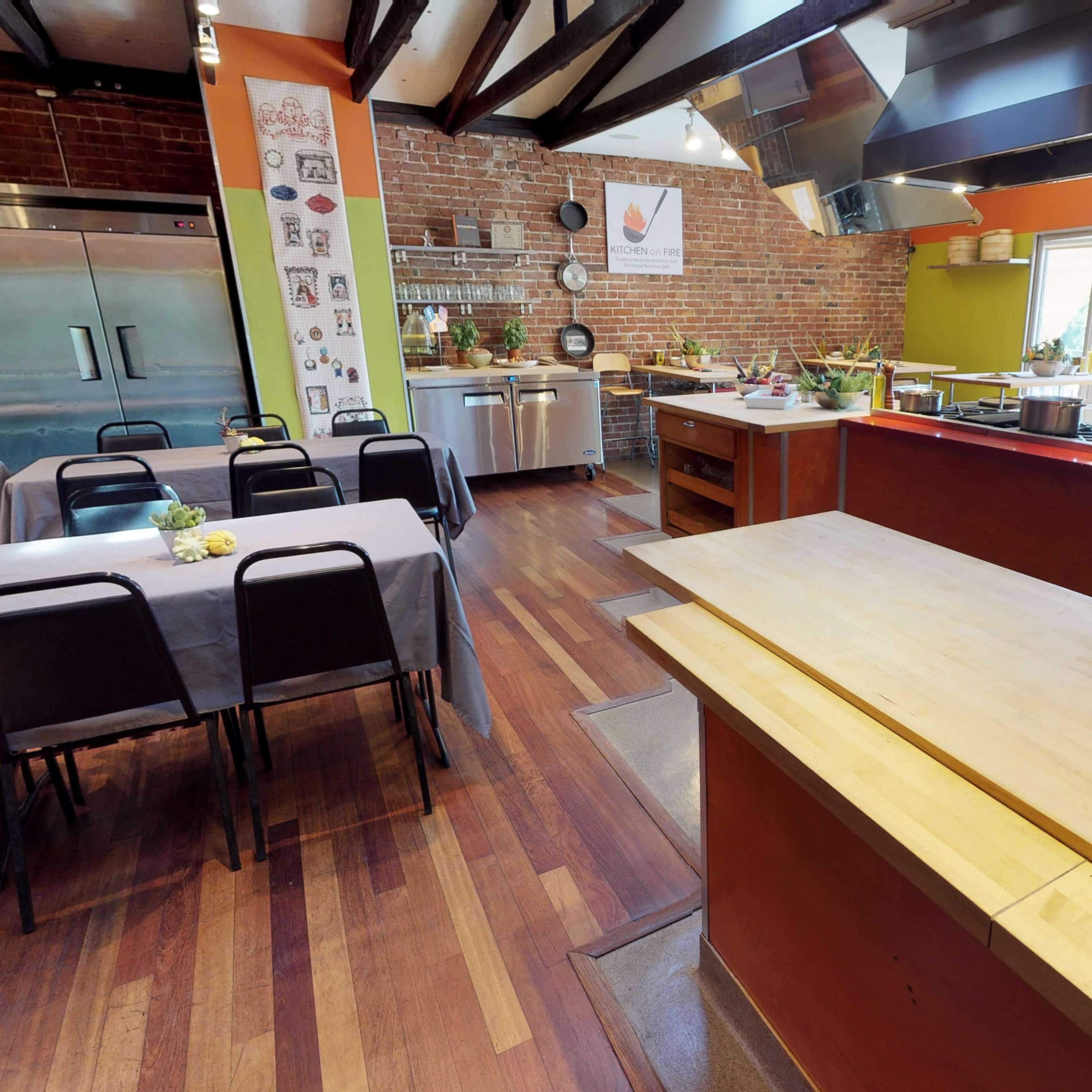 A modern kitchen space with an open layout, featuring wooden floors, stainless steel appliances, and several tables set for cooking classes.
