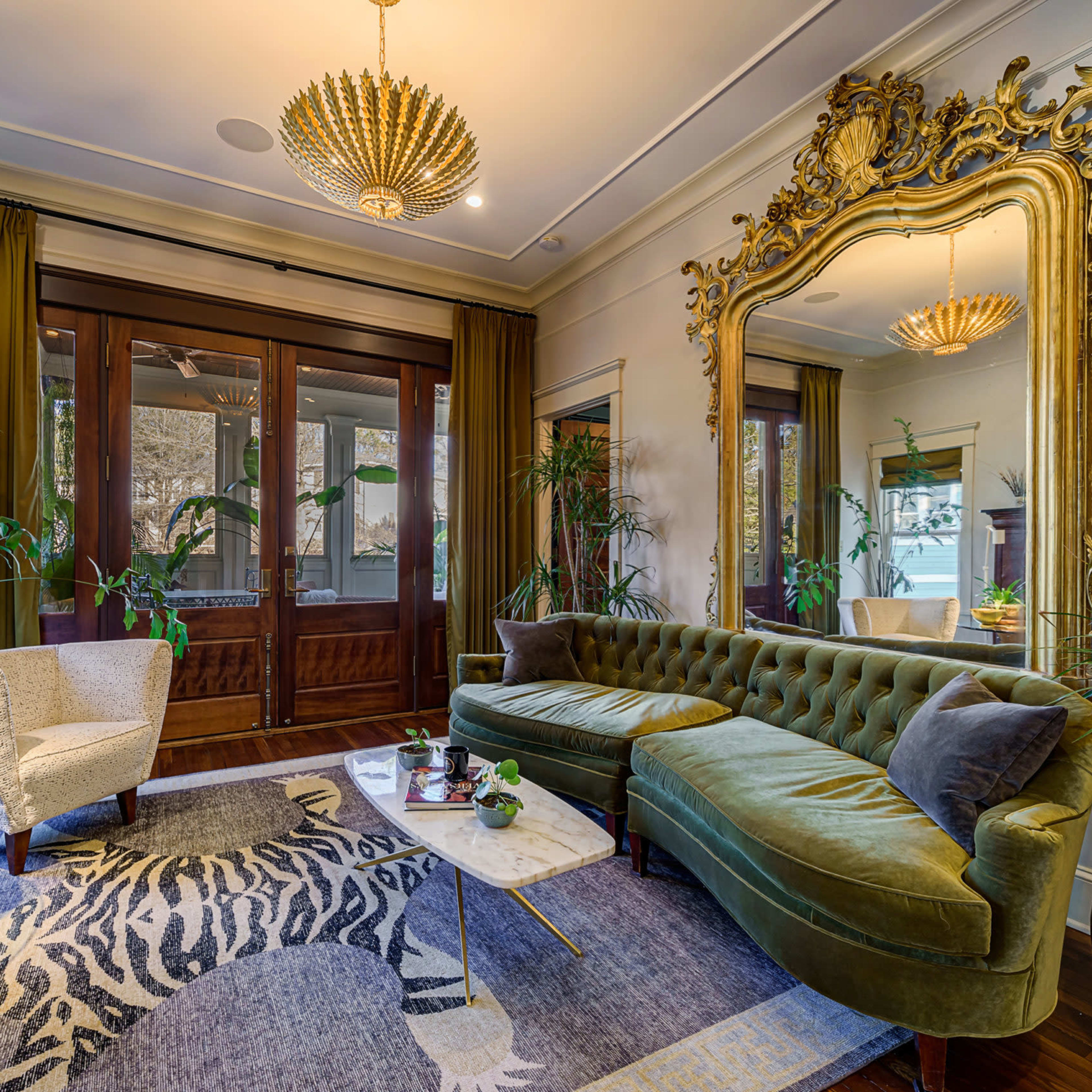 A living room featuring a large green velvet sofa, ornate mirror, potted plants, and a coffee table with a marble top on a patterned rug.