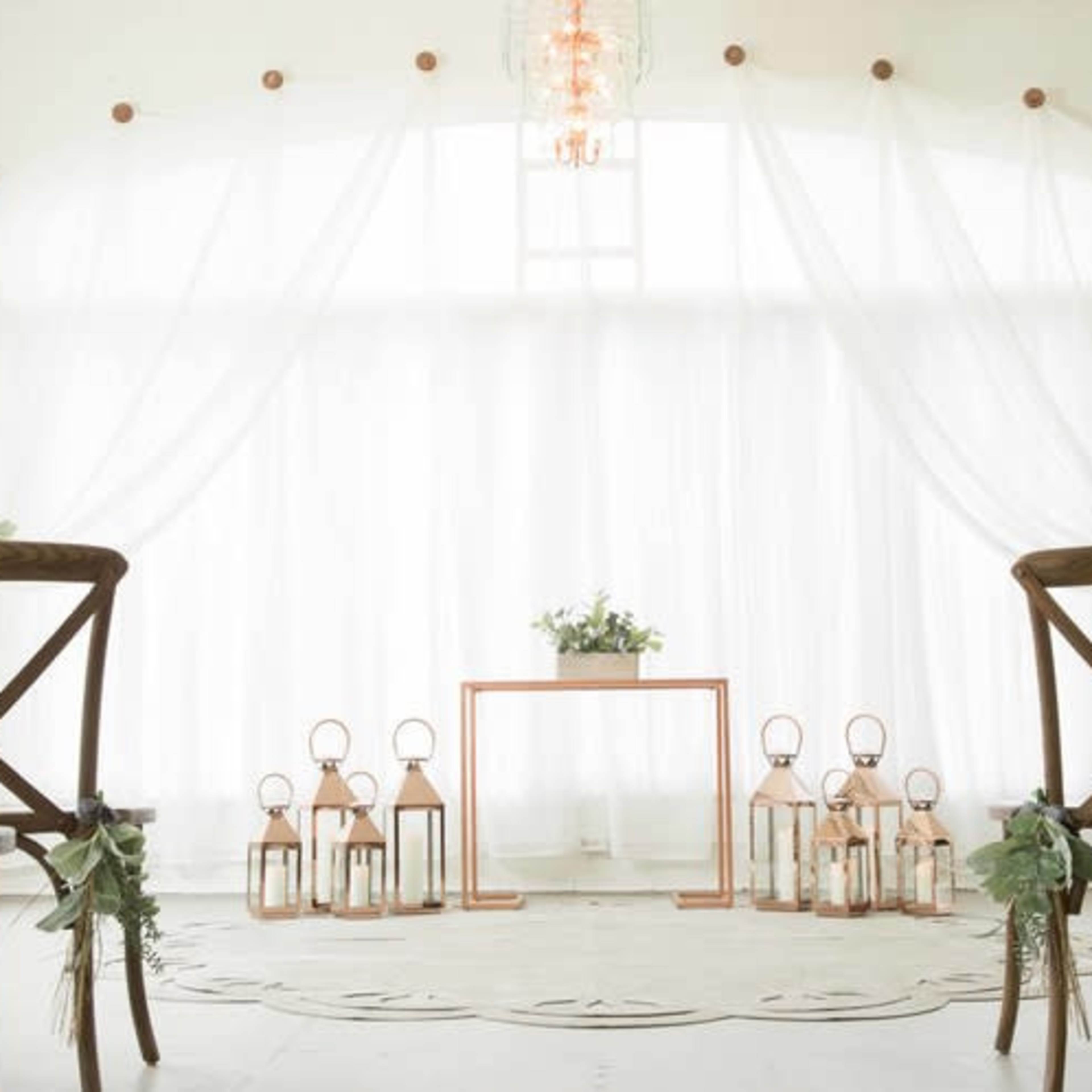 The scene shows a minimalistic wedding setup with wooden chairs facing a small table and decorative lanterns arranged on the floor against a backdrop of draped white fabric.