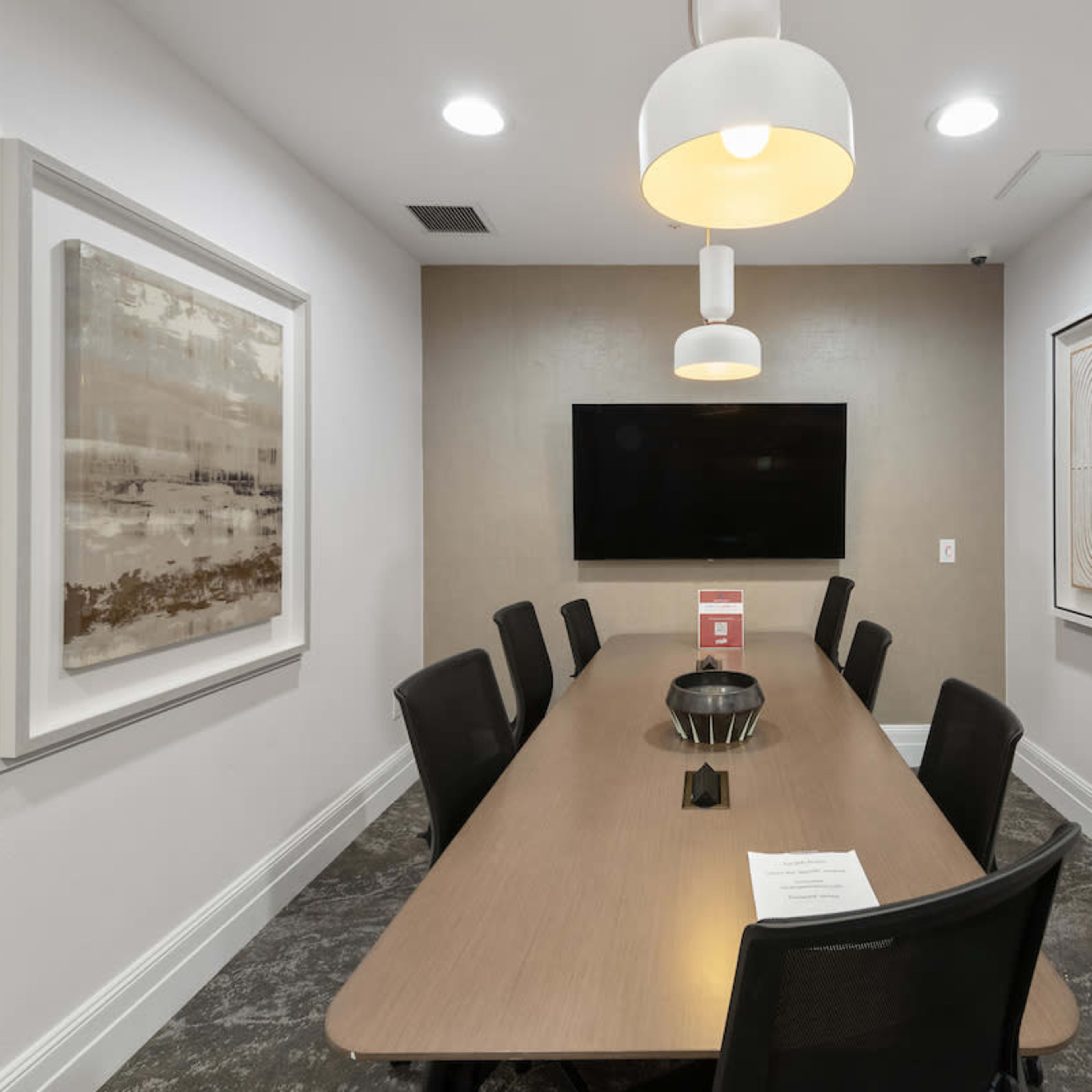 A modern conference room features a long wooden table surrounded by black chairs, with two abstract artworks on the walls and a television mounted at the front.