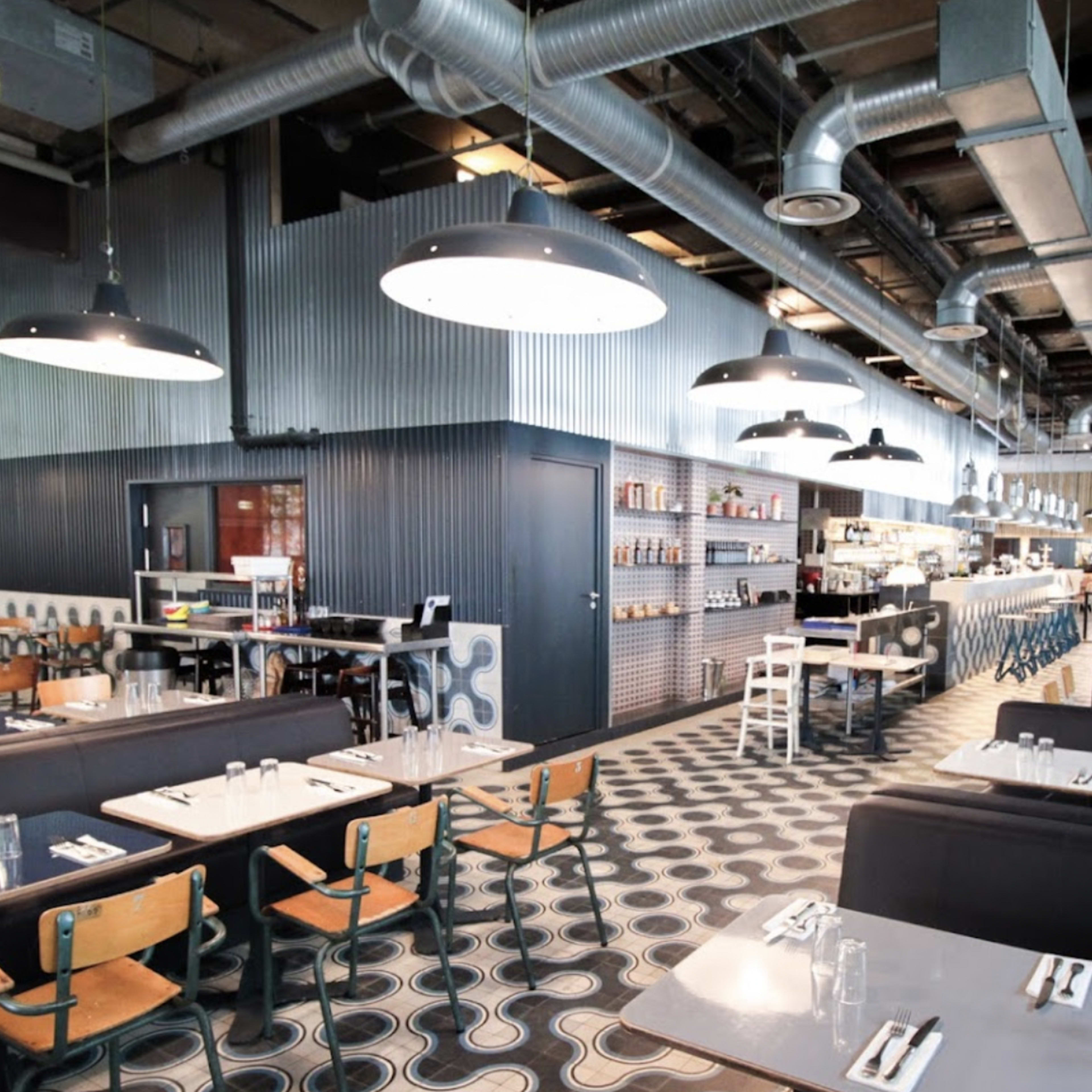 The interior of a modern restaurant featuring an open layout with tables and booths, industrial-style lighting, and a bar area in the background.