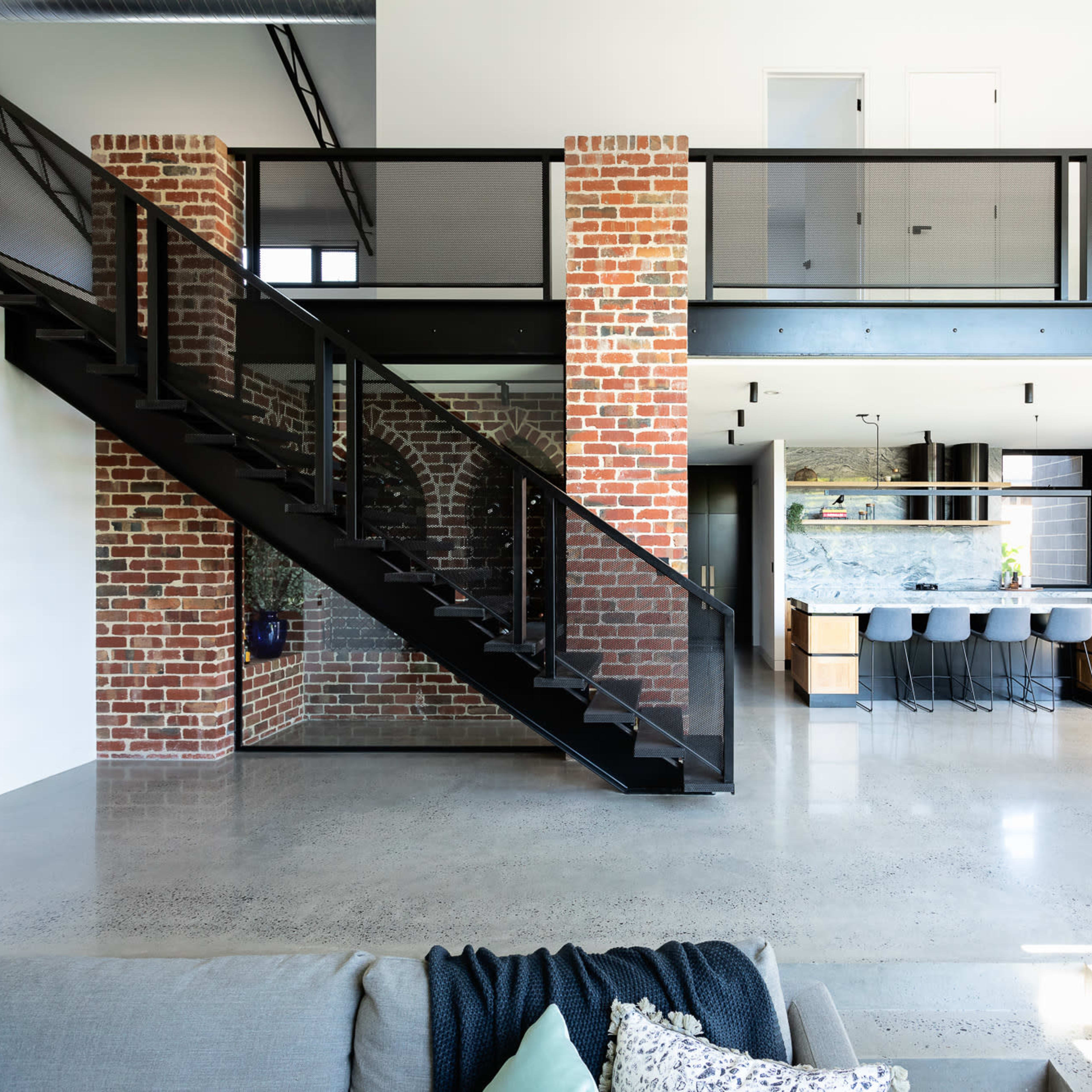 A modern interior with a black metal staircase leading to a mezzanine level, featuring exposed brick walls and an open kitchen and dining area with a large dining table and stools.