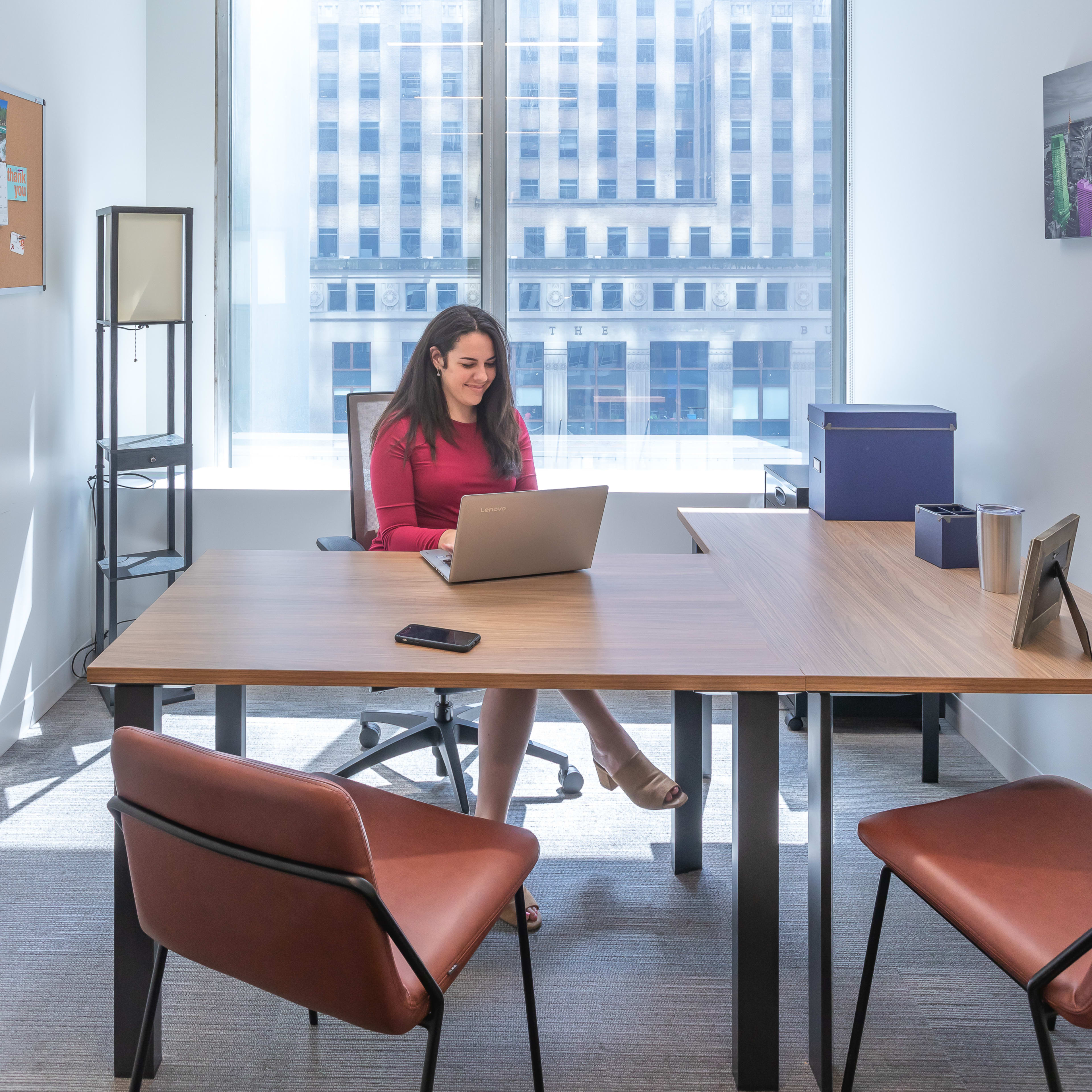 A woman in a red dress is sitting at a wooden desk with a laptop in a well-lit office with large windows.