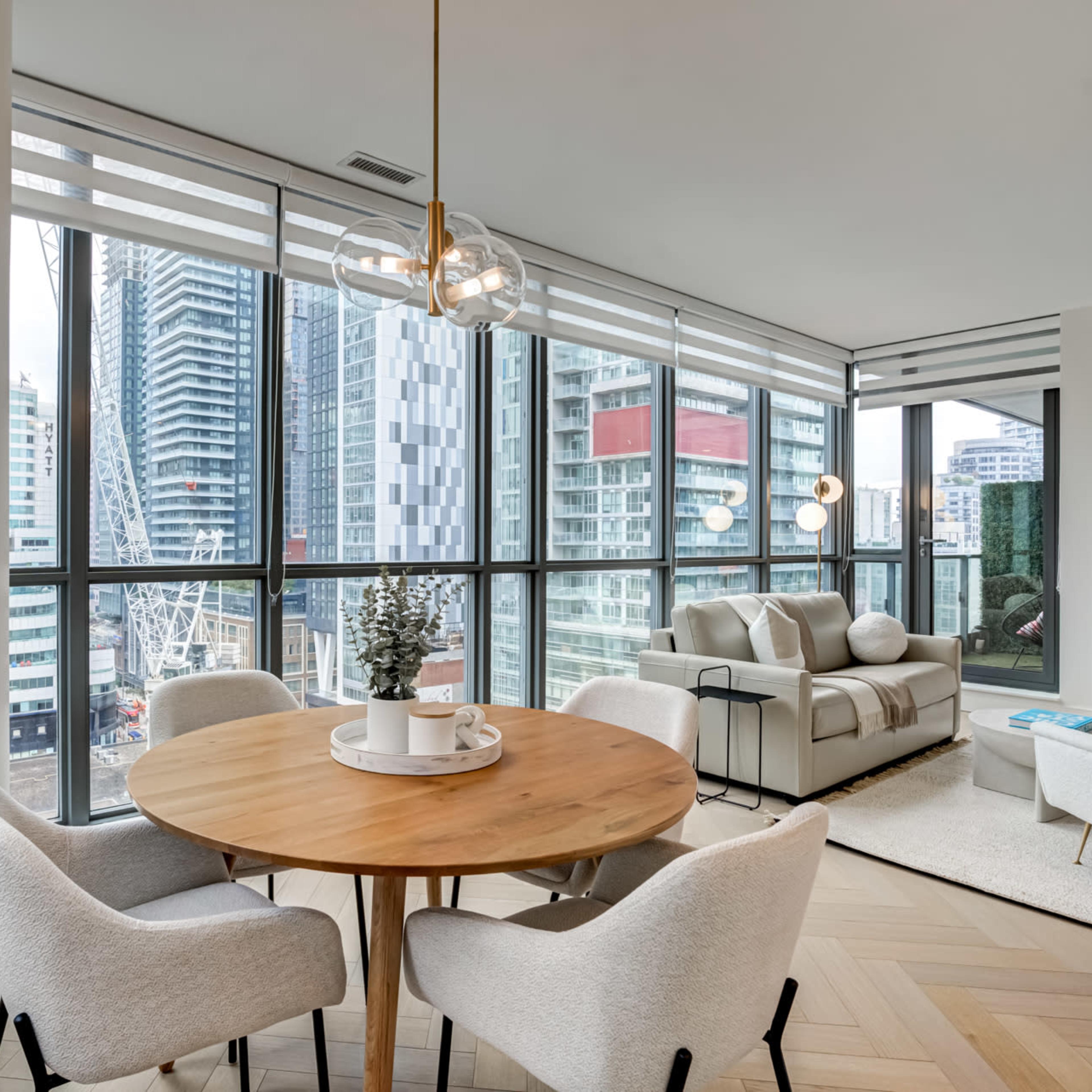 A dining area features a round wooden table surrounded by four upholstered chairs, with large windows showcasing a cityscape and modern furniture in the living space.