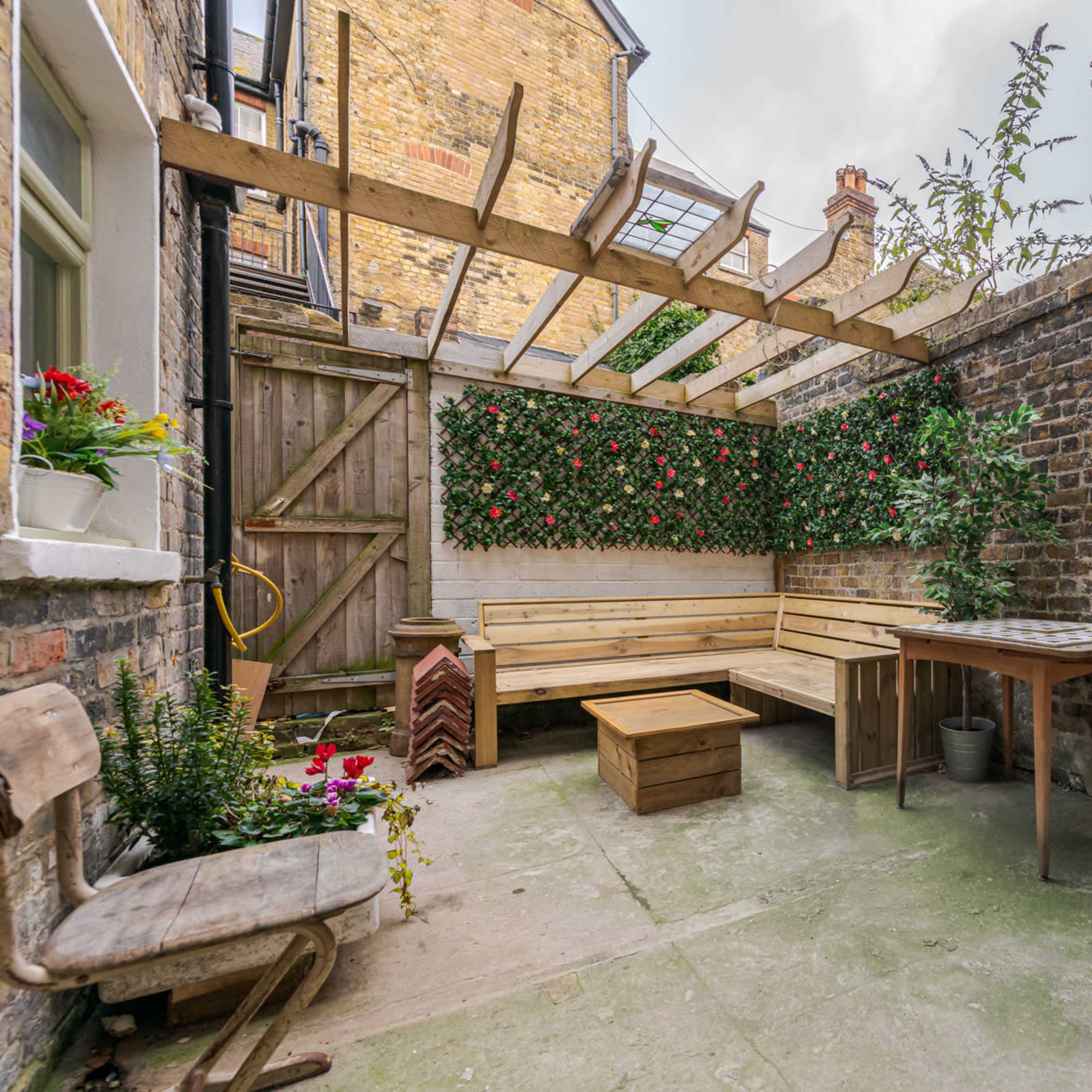 A small, enclosed outdoor courtyard with wooden seating, a table, and a green wall of plants surrounded by brick walls.