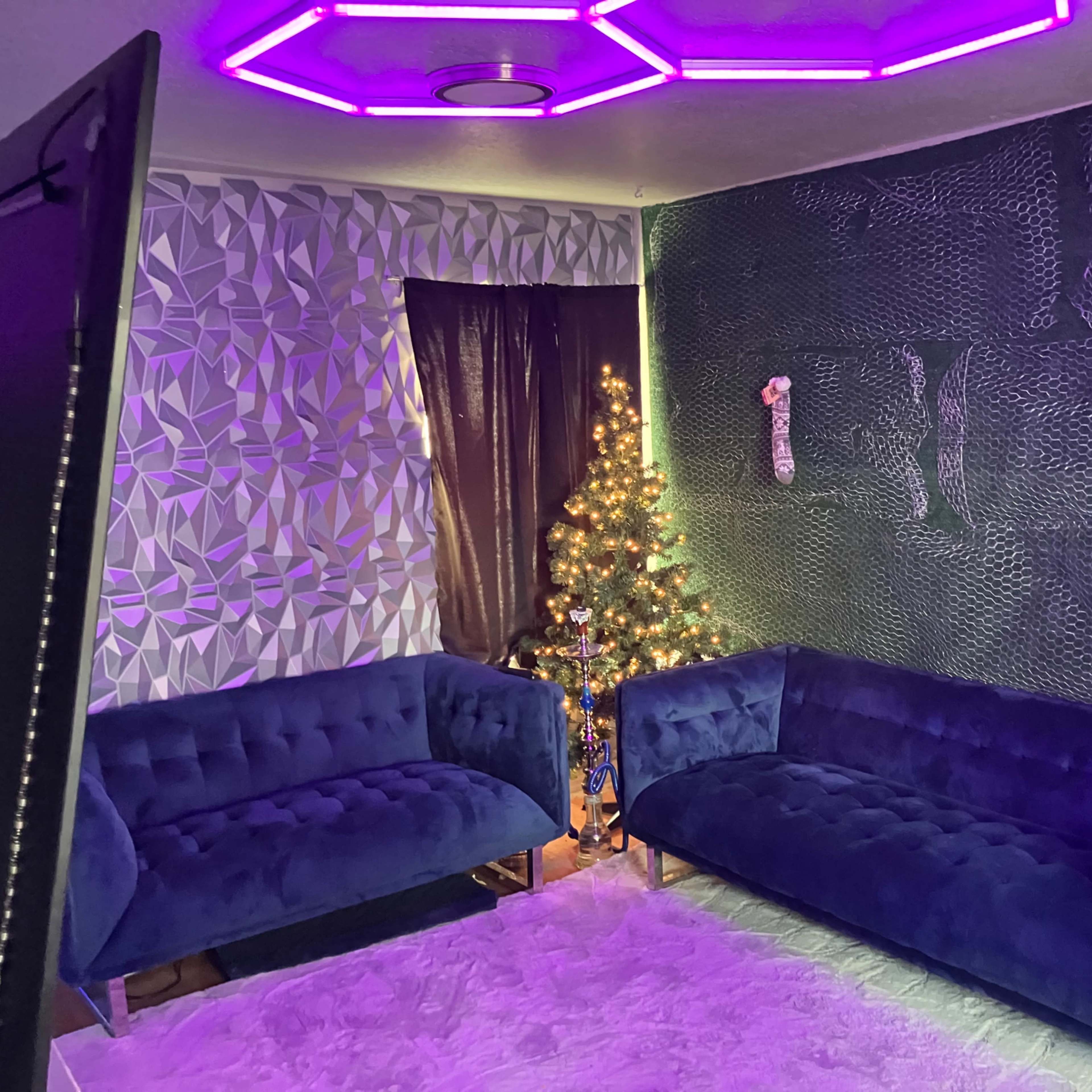 The scene features a modern living room with two blue velvet sofas, a decorated Christmas tree, and a wall adorned with patterned wallpaper and purple LED lighting.