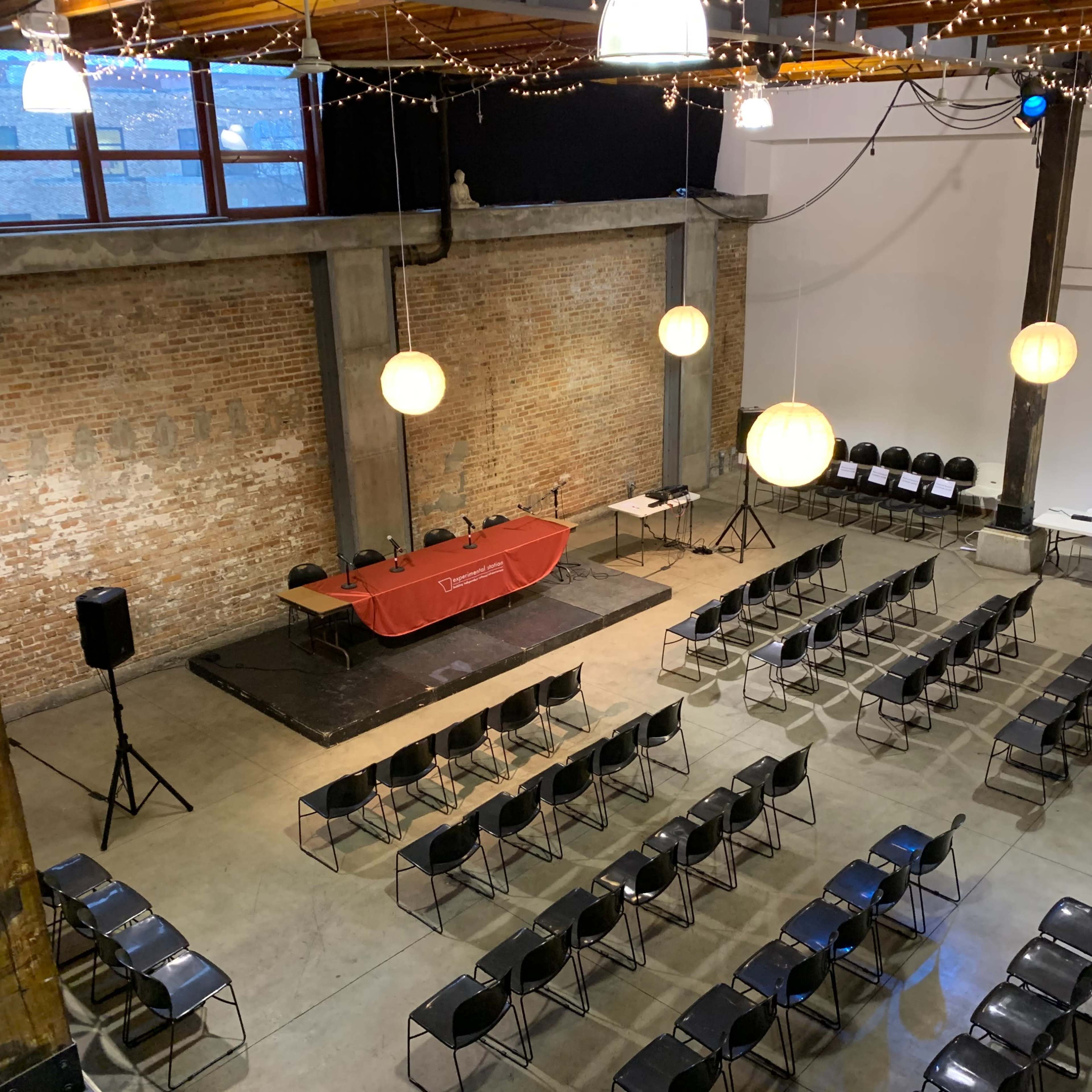 An empty event space with rows of black chairs facing a red table set up on a stage, accompanied by hanging light fixtures and exposed brick walls.