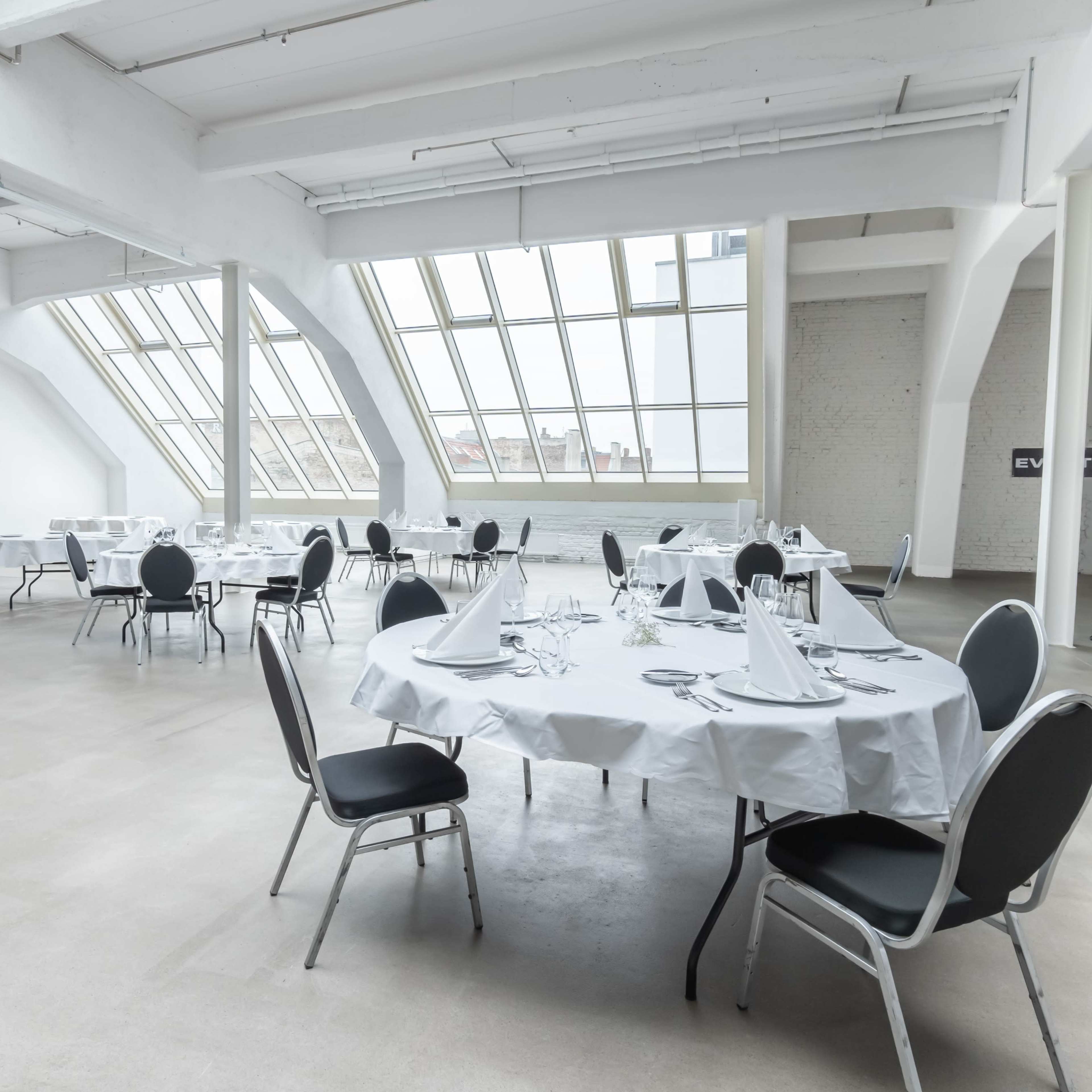 A spacious, modern event space features neatly arranged round tables with white tablecloths and chairs, illuminated by large windows.