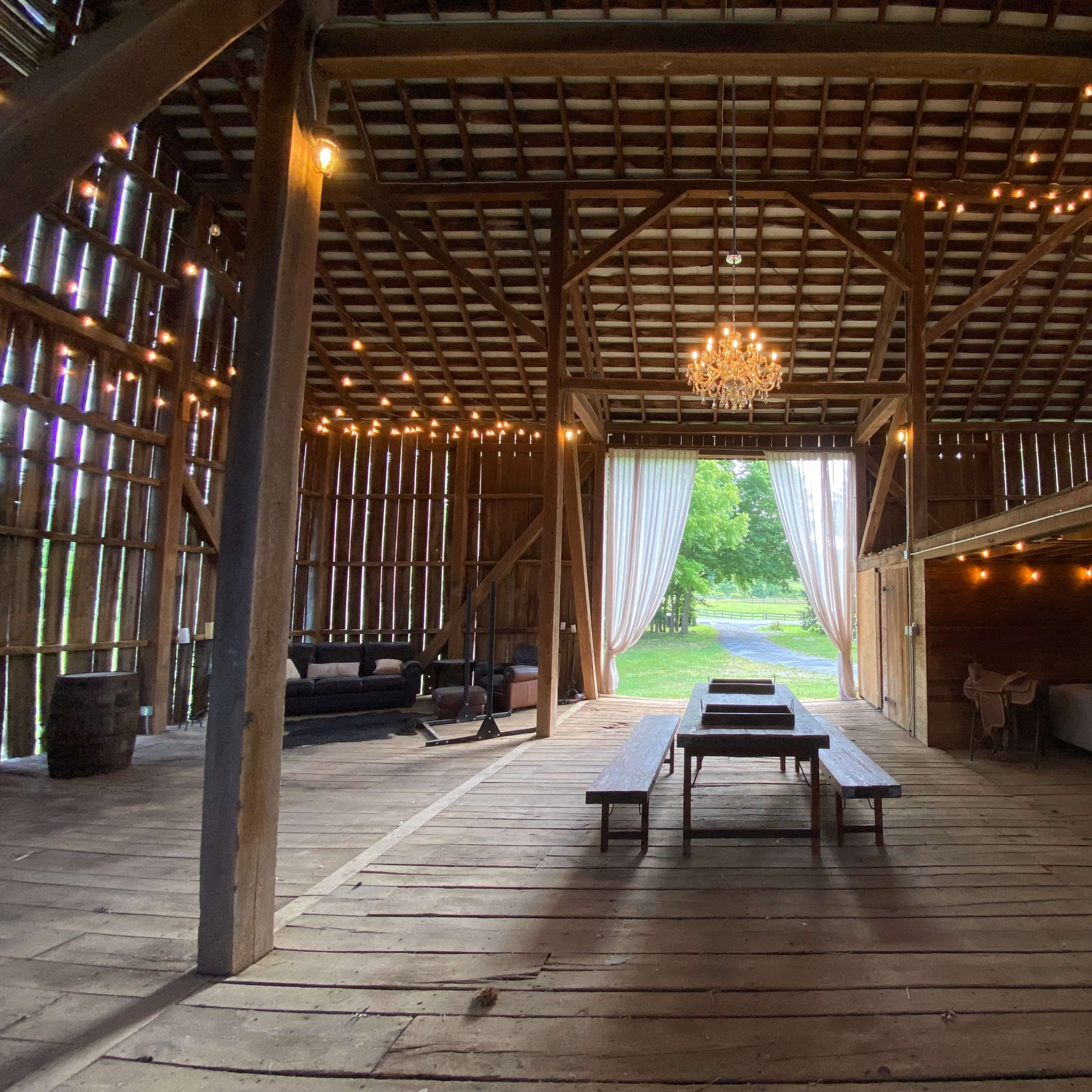 The interior of a rustic barn featuring wooden floors, string lights, a chandelier, and furniture arranged around a central table.