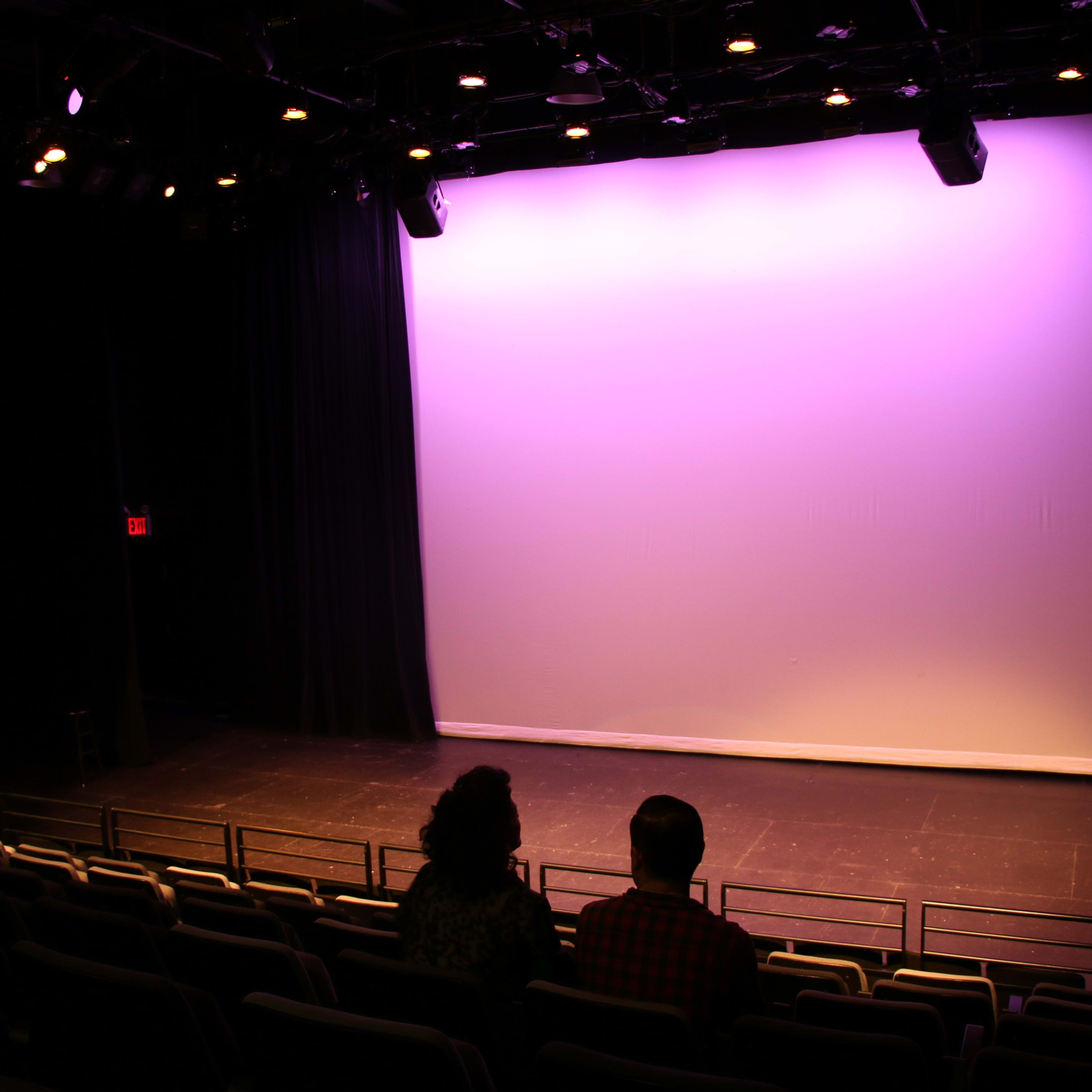 A couple sits in an empty theater with a large purple-lit stage in front of them.