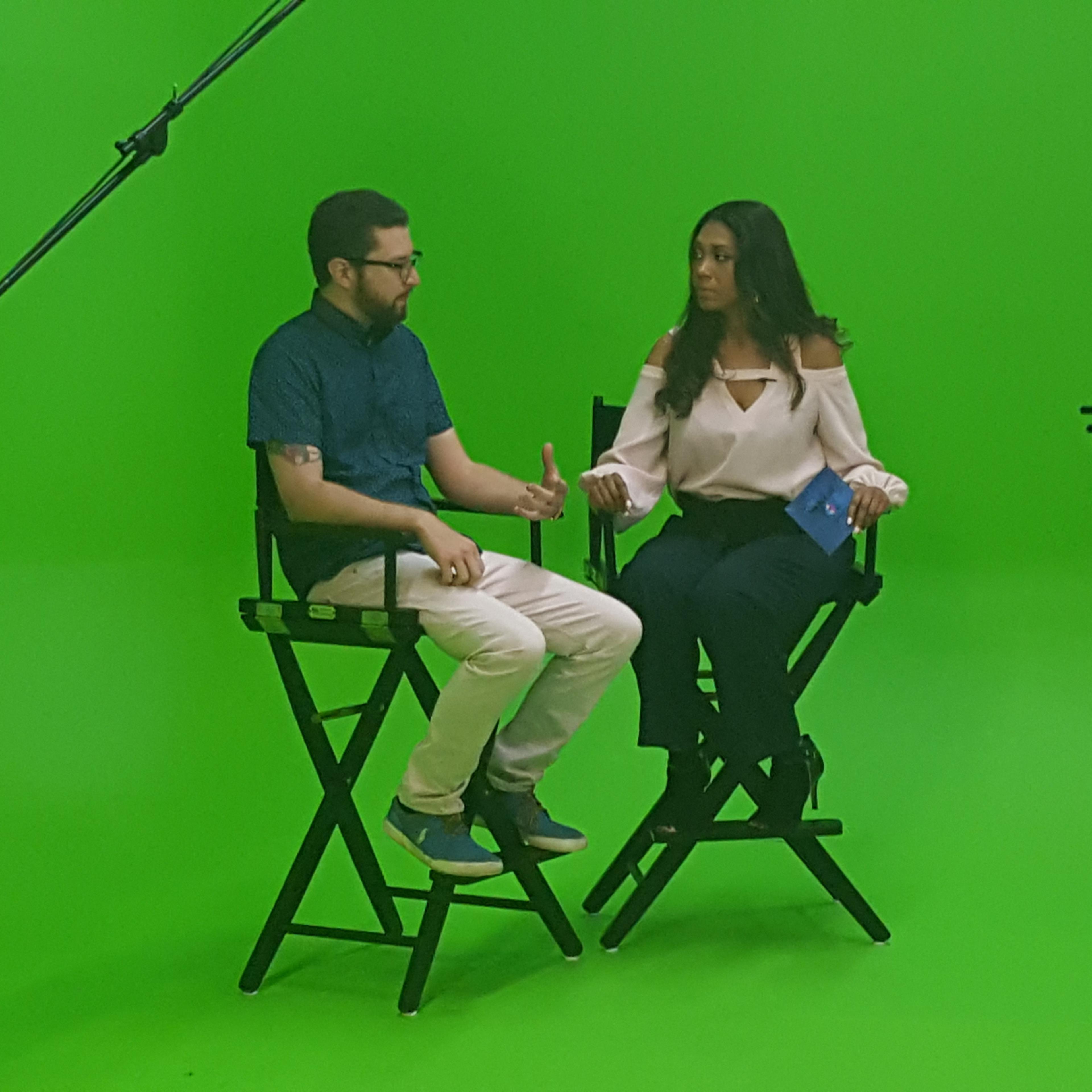 A man and a woman sit in director's chairs on a green screen set while a camera operator records the scene.