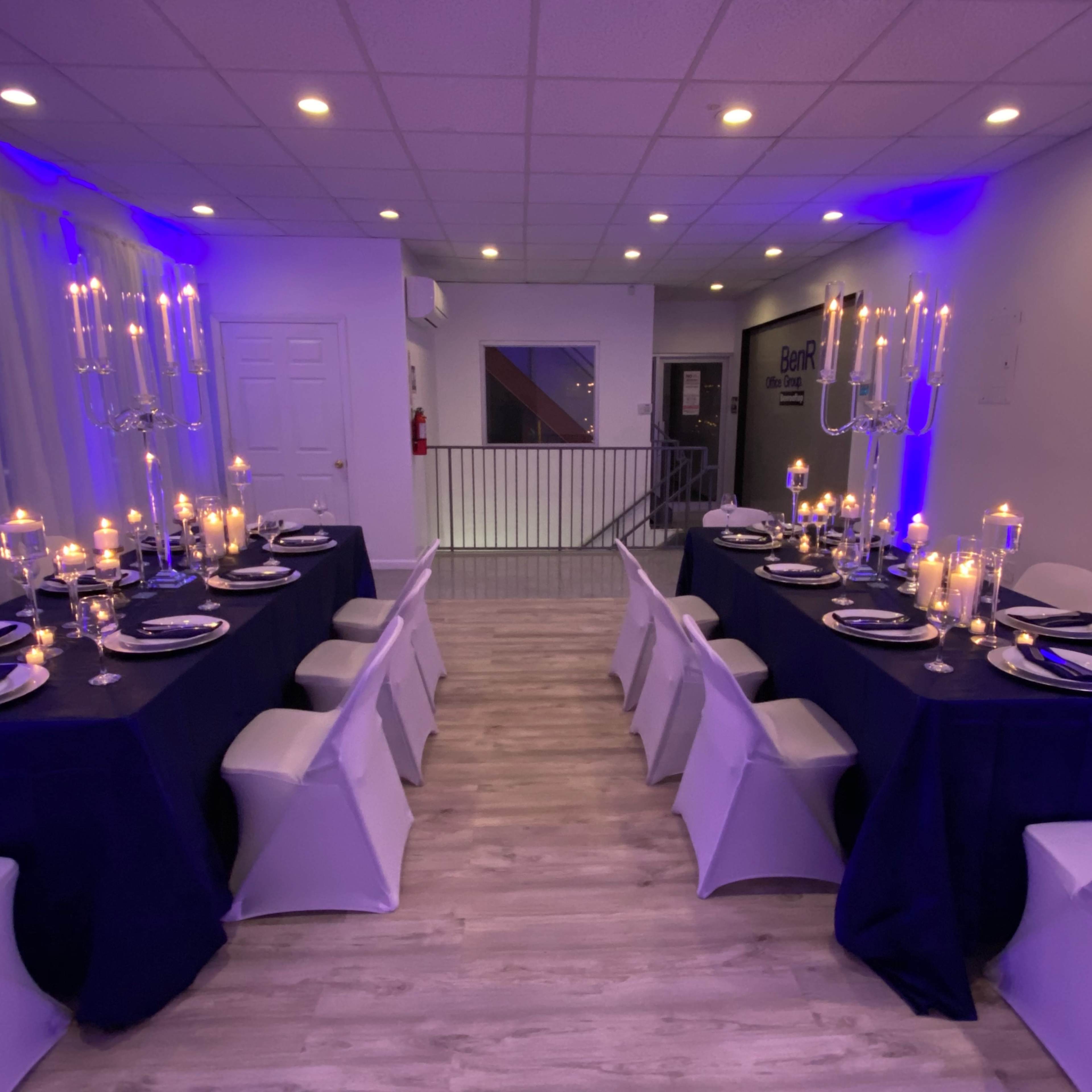 A dining room set up with two long tables adorned with dark tablecloths, plates, candles, and elegant seating arrangements under blue lighting.