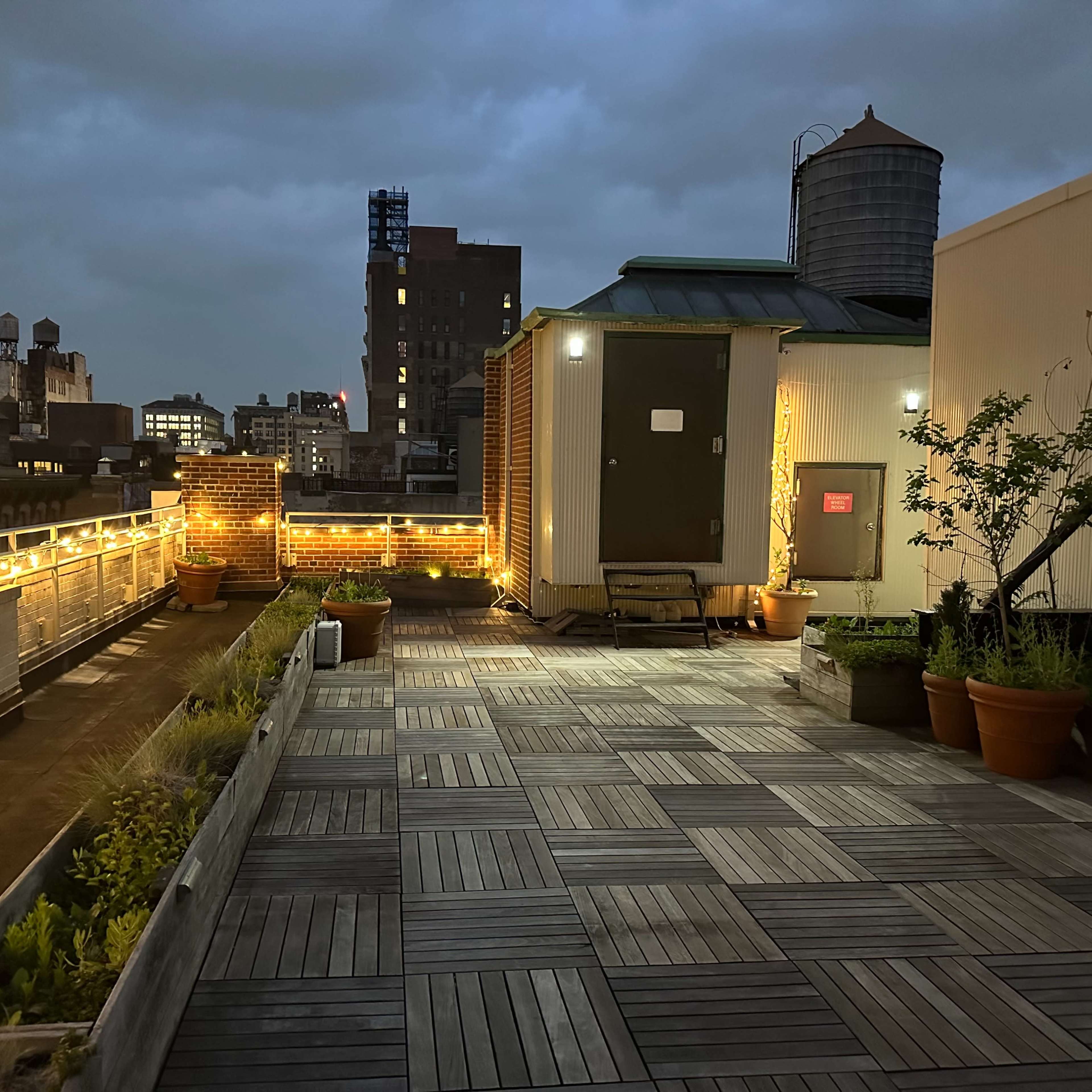 A rooftop terrace with wooden flooring, planters, and soft lighting against a city skyline at dusk.