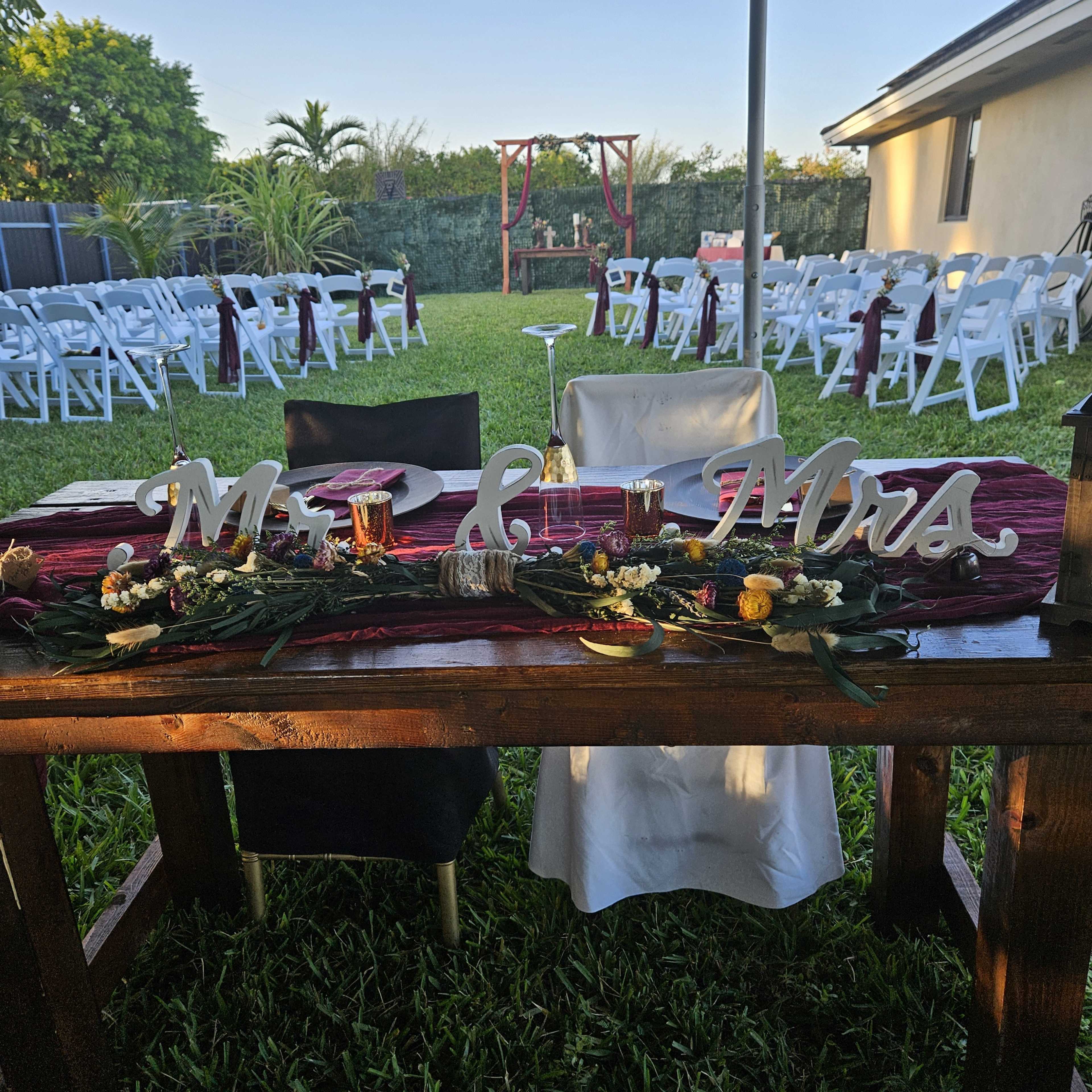 A decorated wedding table with "Mr. & Mrs." letters sits in a grassy area, surrounded by chairs set up for guests.