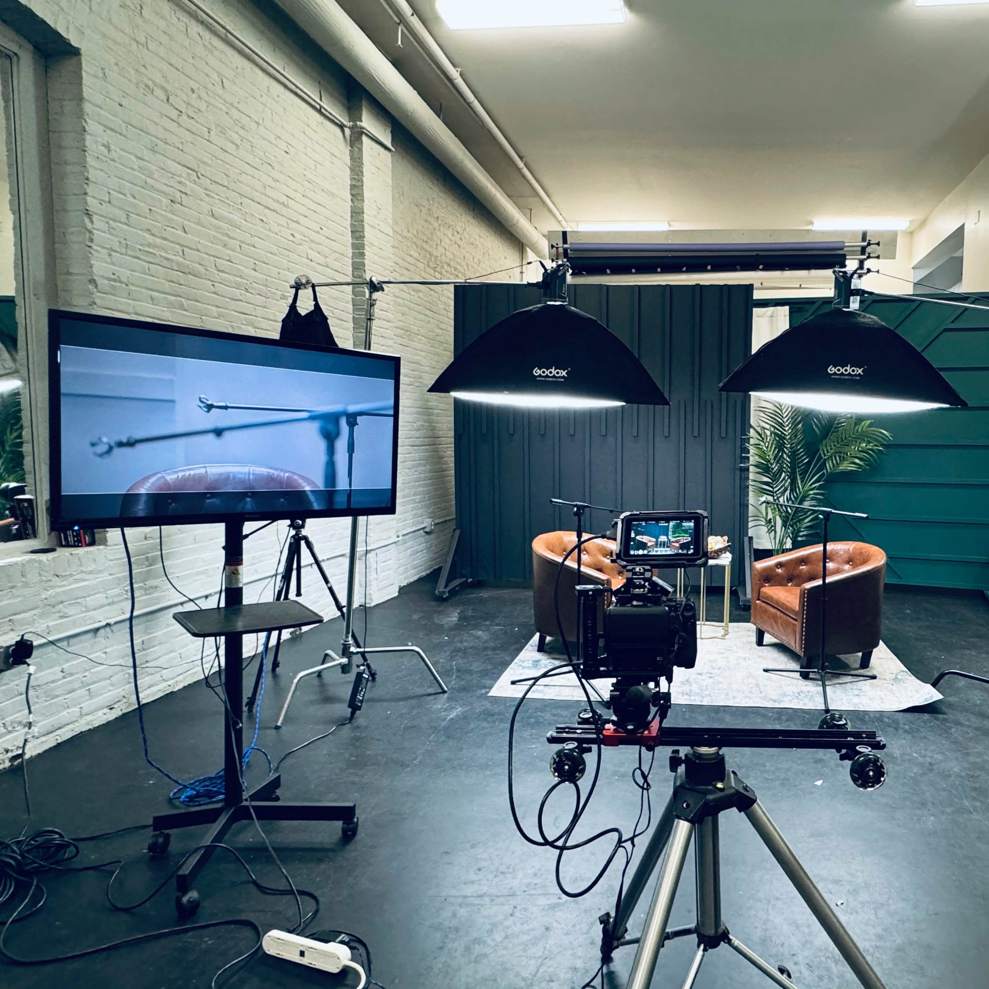 A video recording setup featuring two brown chairs, various cameras, and lighting equipment arranged in a studio space.