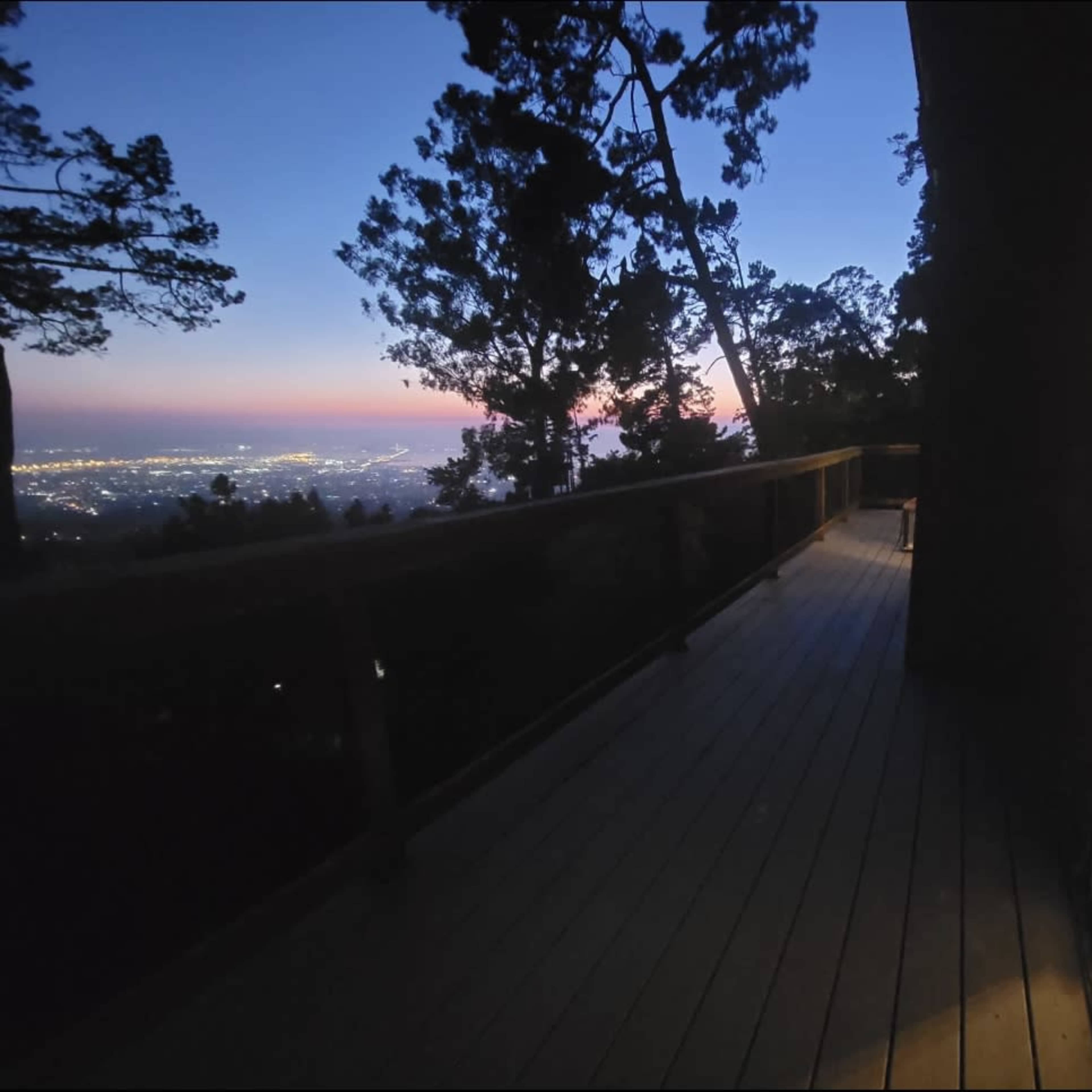 A wooden deck overlooks a city lit at dusk, with a small interior space visible through an open sliding door.