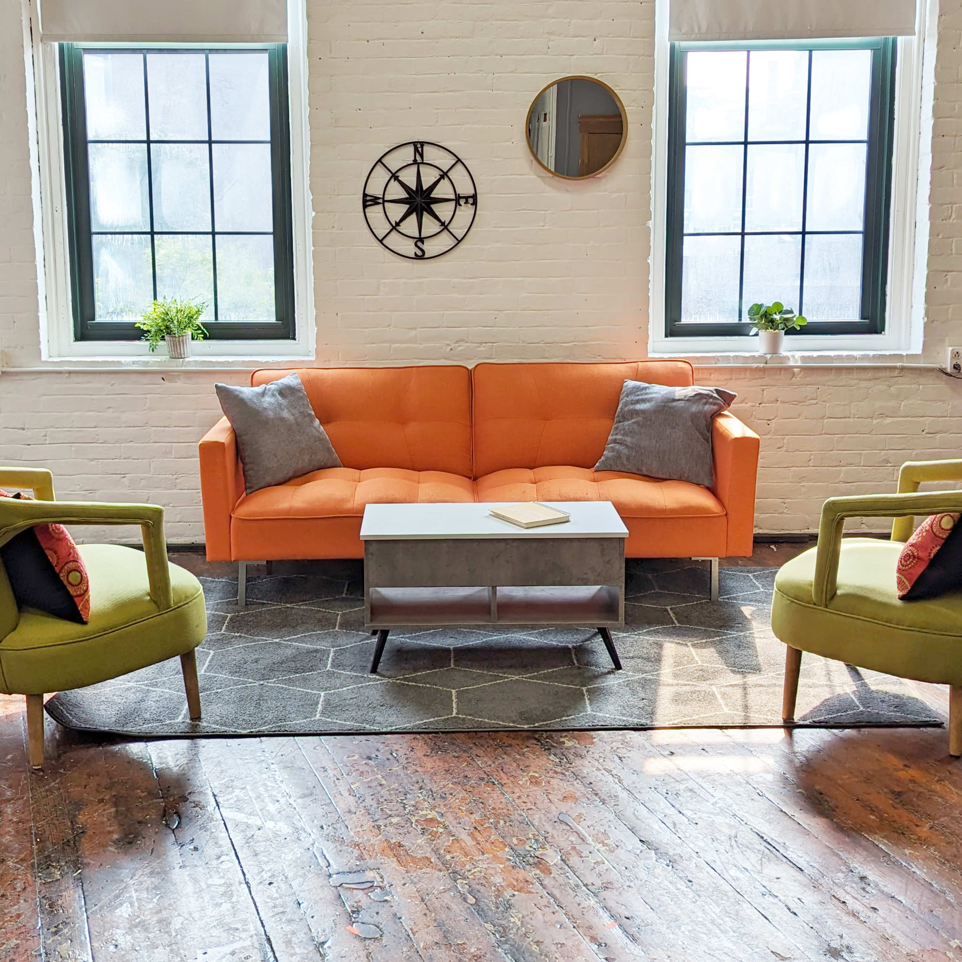 A cozy living room with an orange sofa, two green armchairs, a coffee table on a hexagonal rug, and large windows letting in natural light.