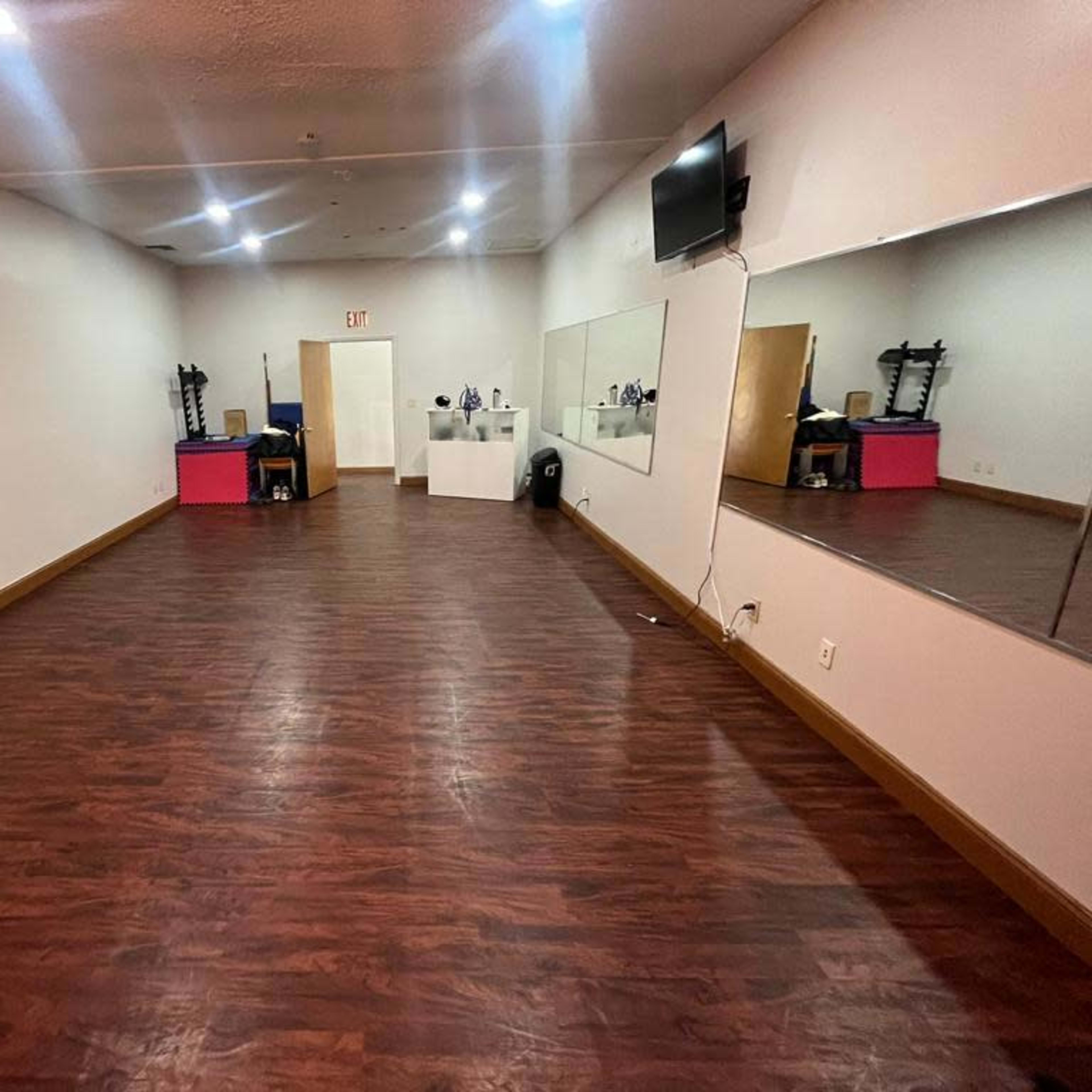 A spacious, empty room with wooden flooring, a large wall mirror, exercise equipment, and a wall-mounted television.