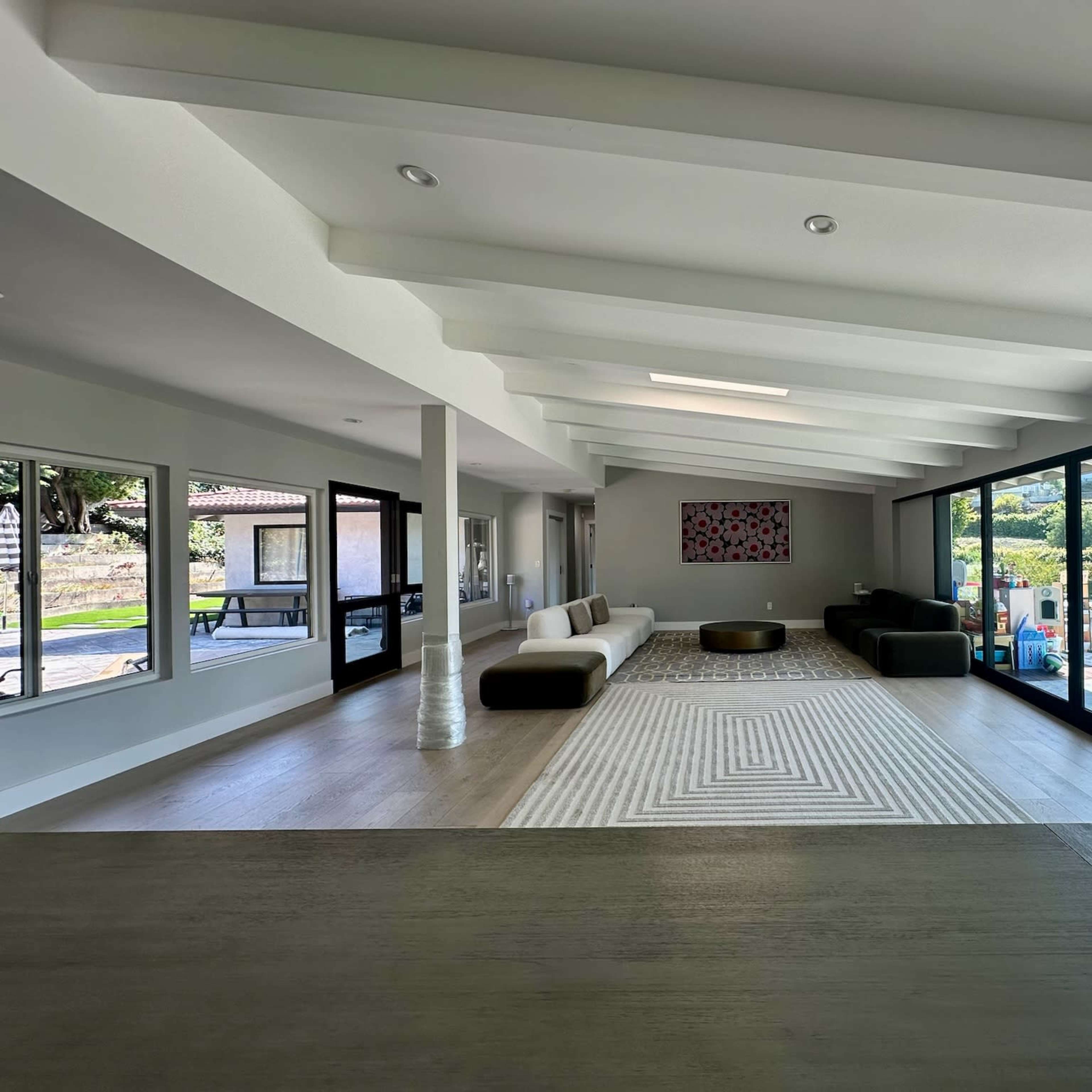 A spacious living room features large windows, a patterned rug, and modern furnishings overlooking a landscaped exterior.