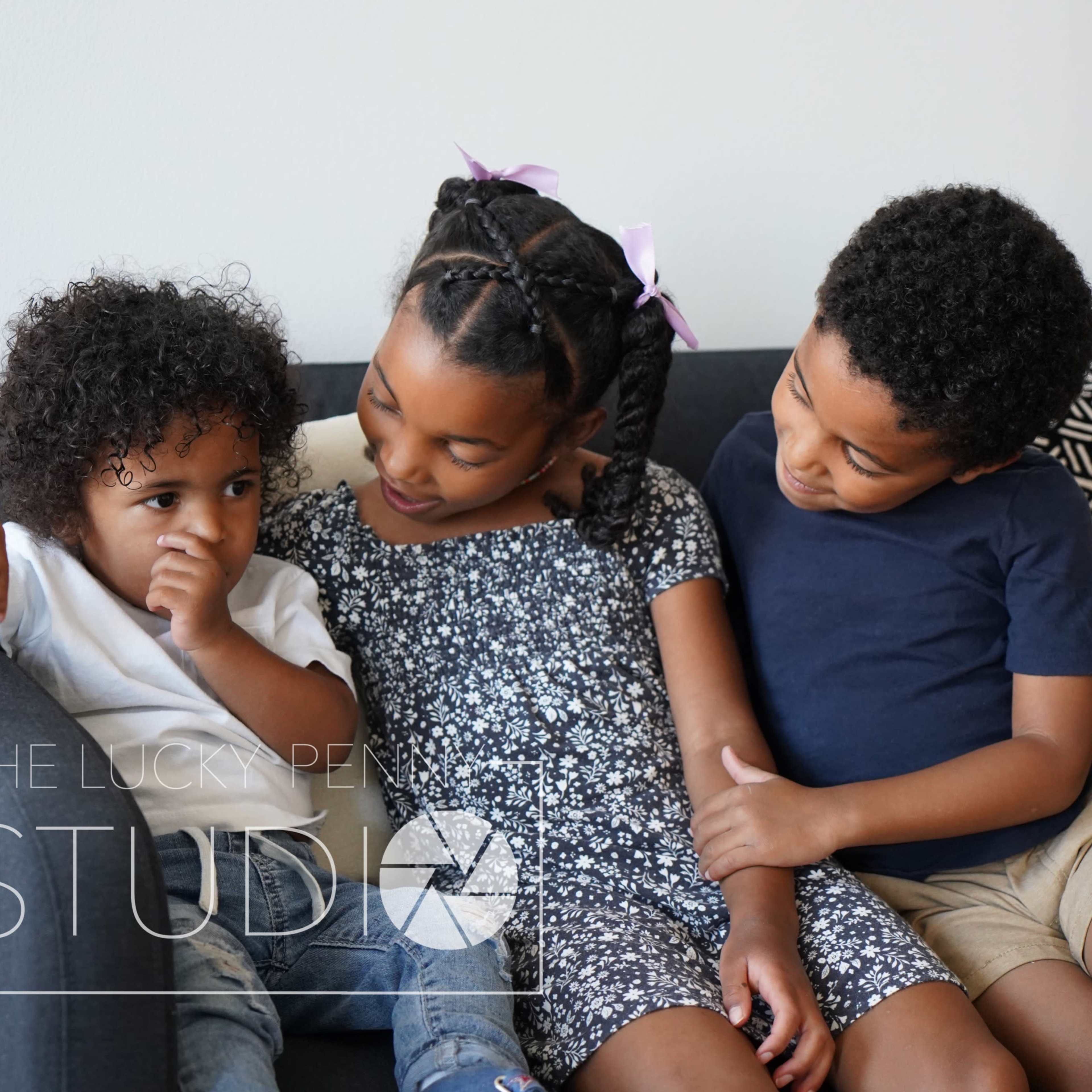 Three children sit together on a couch, with one child resting their finger on their mouth while the others look at them.