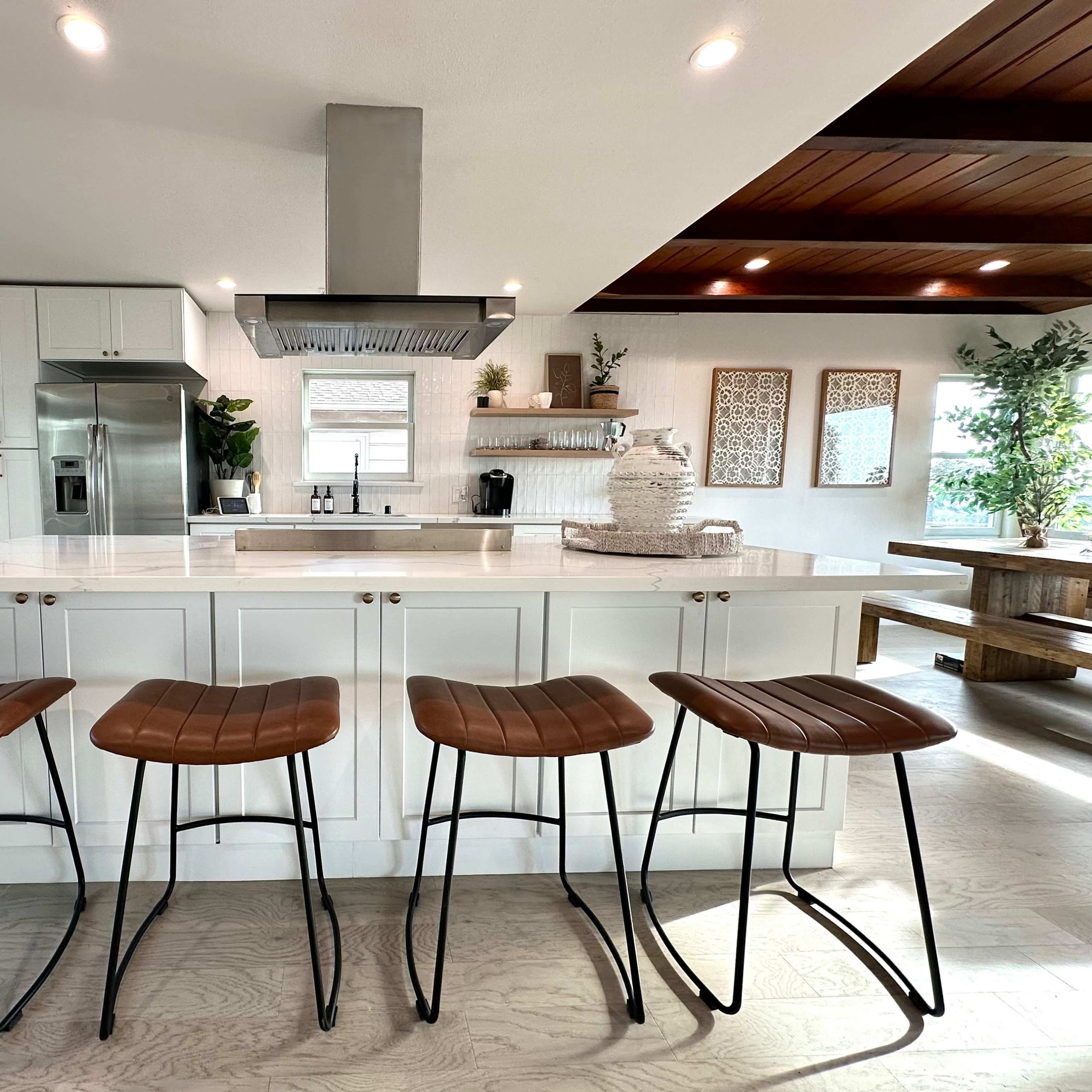 A modern kitchen with a central island featuring four metal-framed, wooden seating stools and stainless steel appliances.