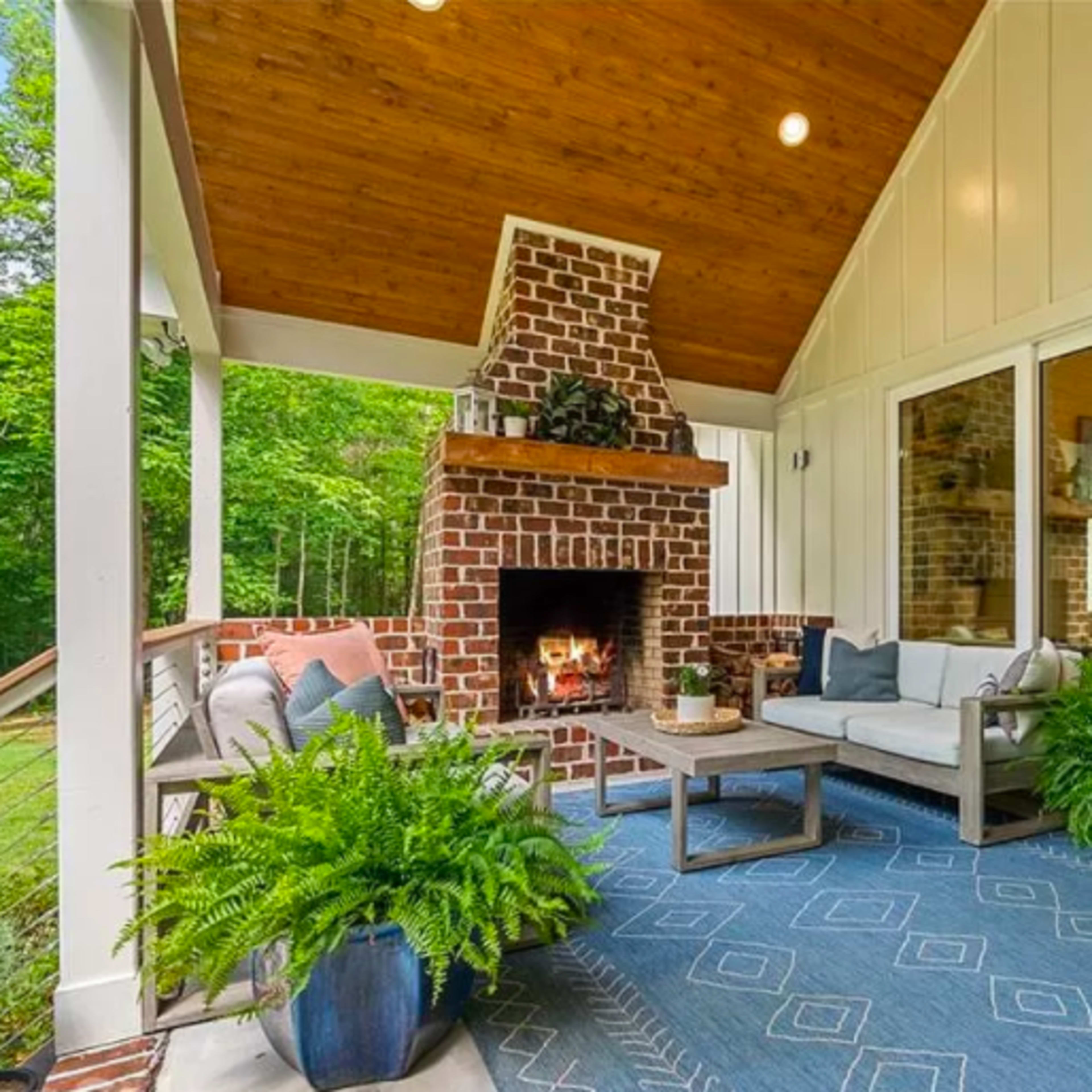 A cozy outdoor living area featuring a brick fireplace, comfortable seating, and large windows overlooking a green landscape.