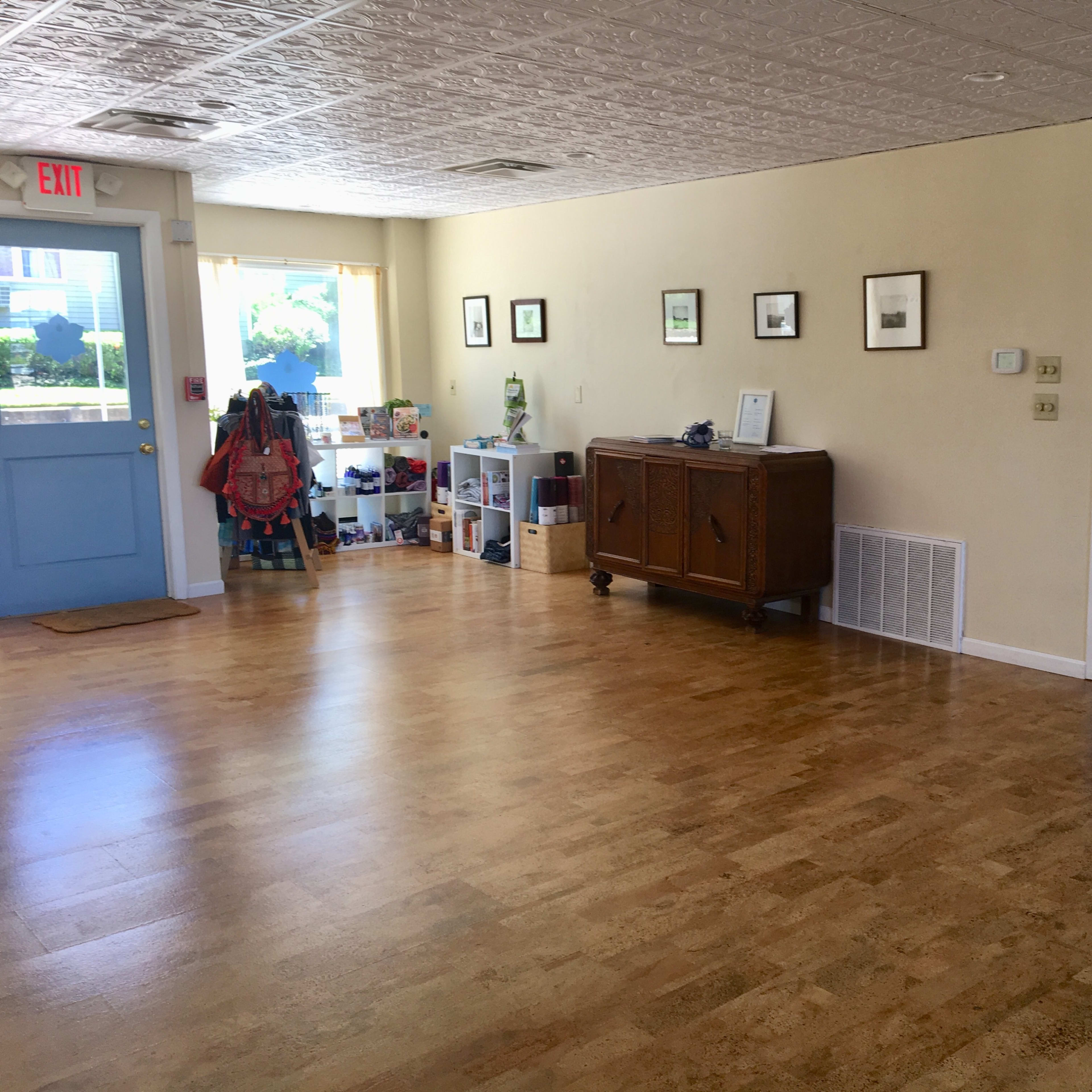 A spacious room with light-colored walls, a wooden floor, and a blue door leading to the outside, along with various framed pictures on the walls and a side table near the entrance.