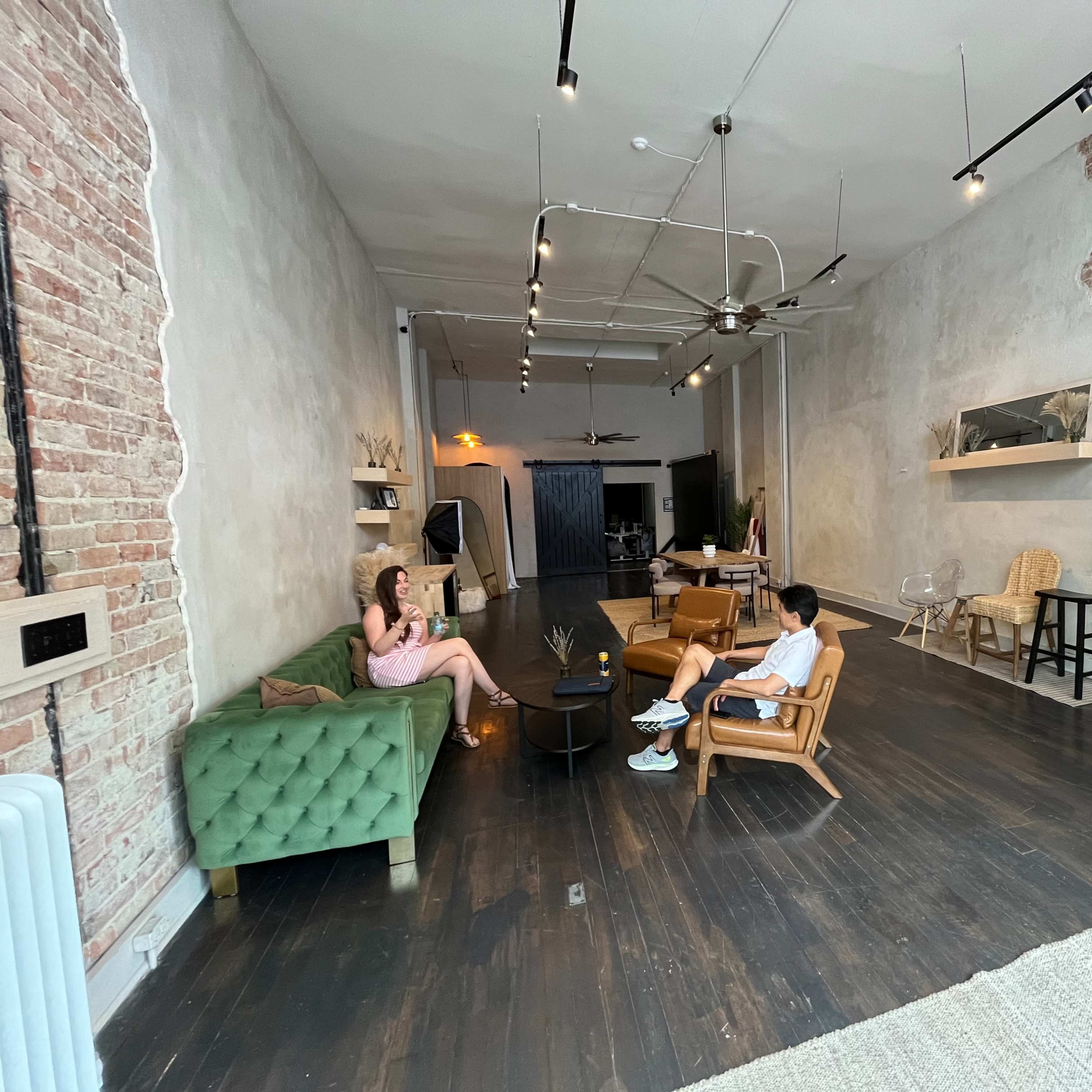 The interior of a modern, industrial-style room features a green velvet sofa, wooden chairs, and exposed brick walls, with two people seated and engaged in conversation.
