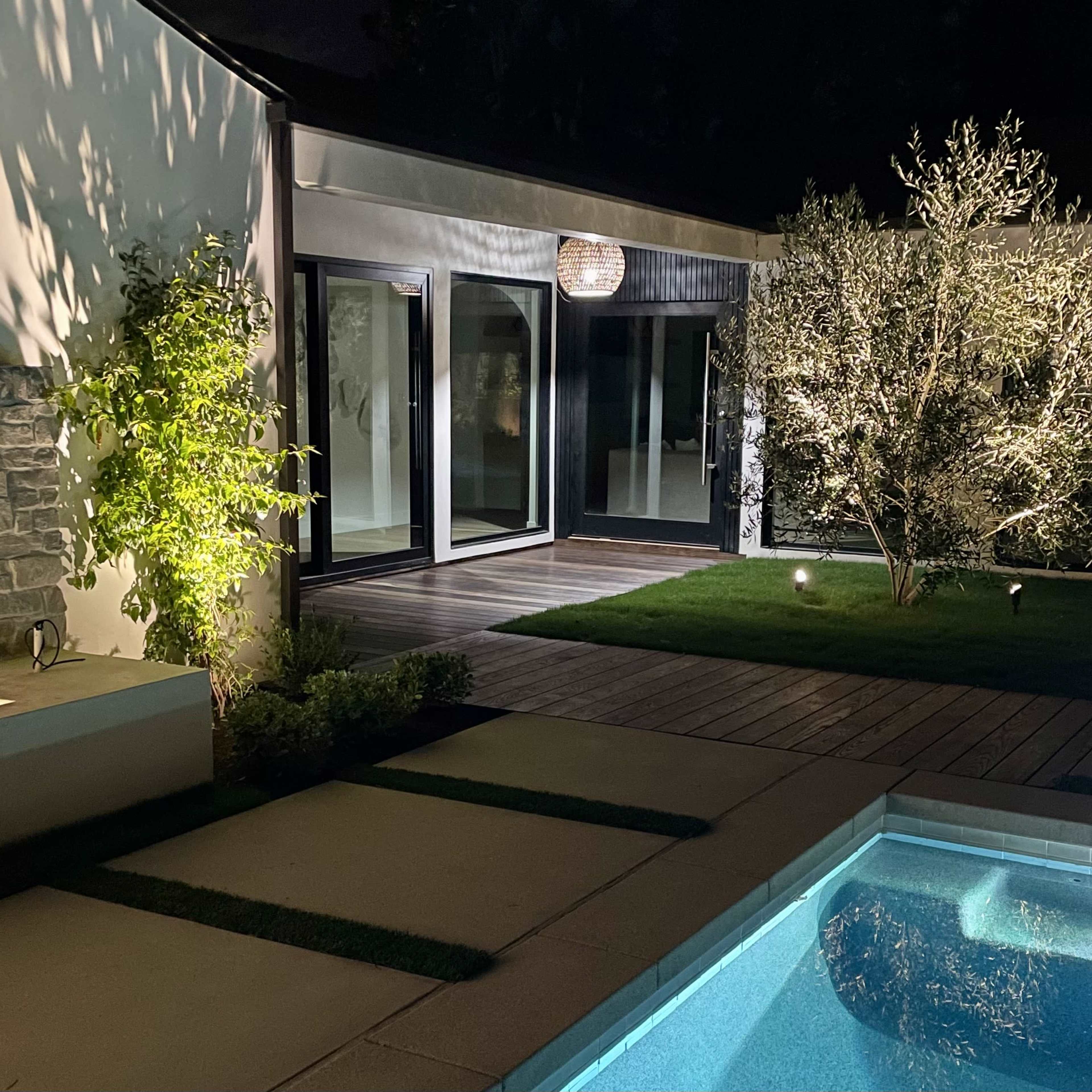 A modern outdoor area at night with a swimming pool, a landscaped garden featuring an olive tree, and illuminated elements near a stone wall.