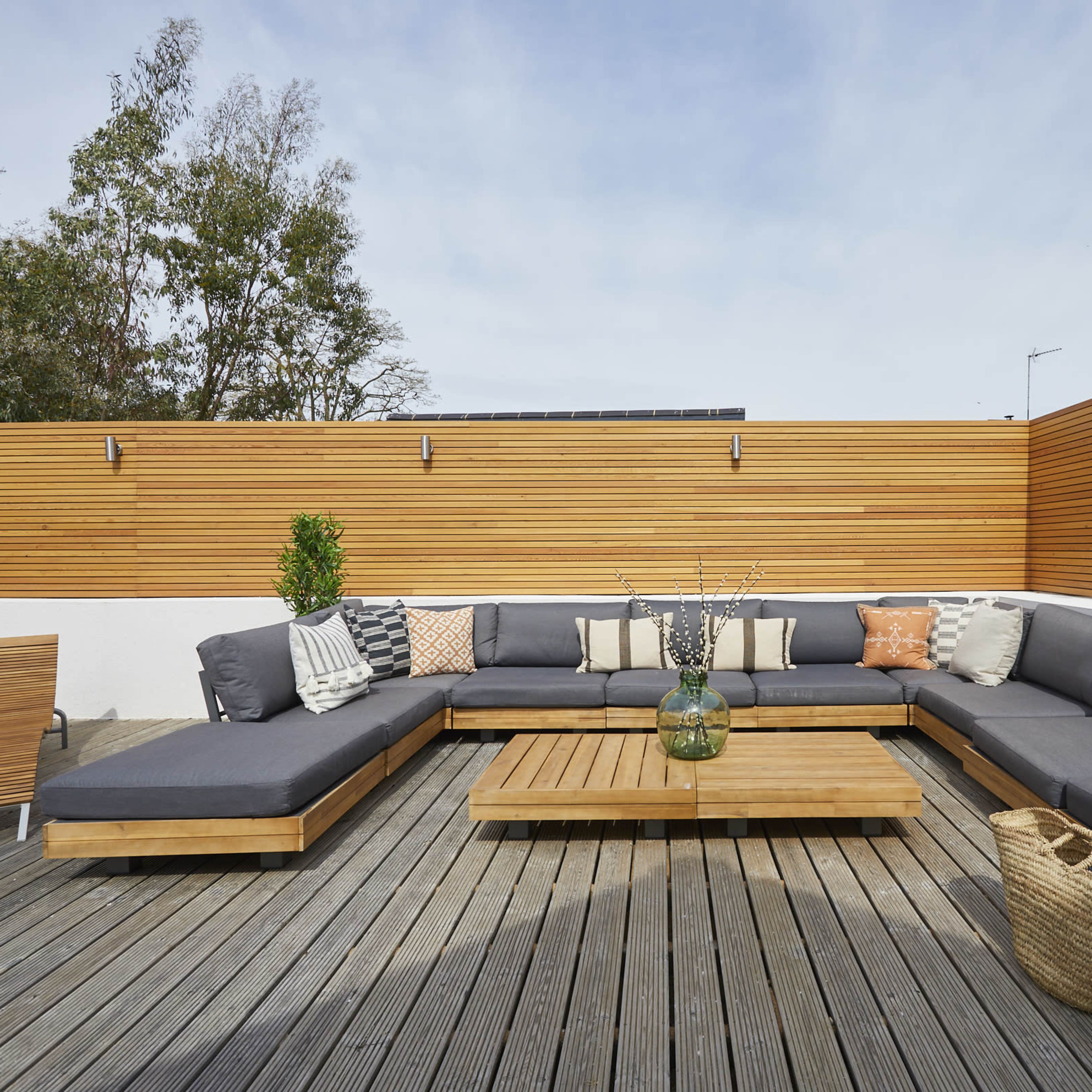 A modern rooftop terrace features a large sectional sofa, a wooden coffee table, and decorative plants against wooden paneling.