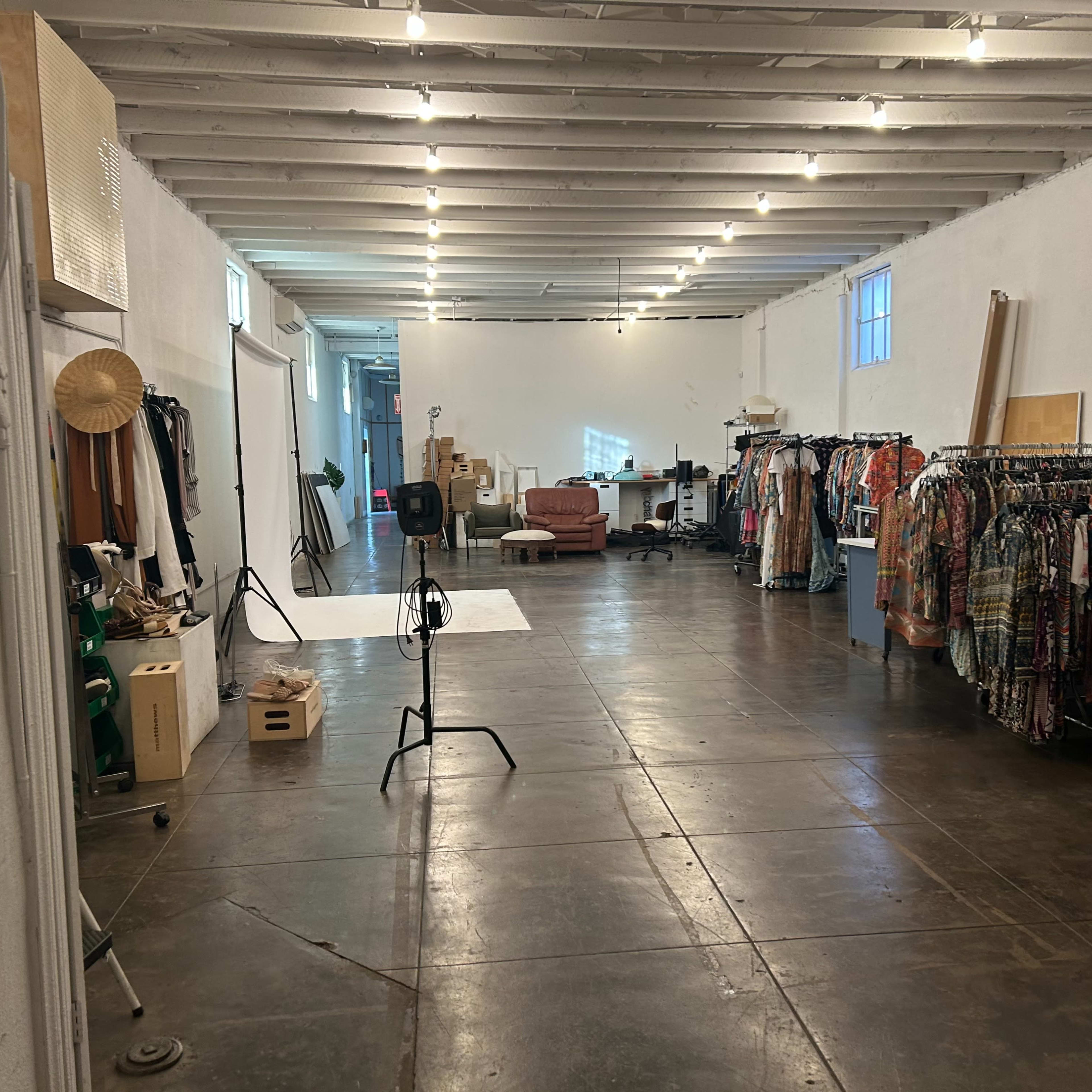 A spacious room with clothing racks filled with various garments, a white backdrop set up for photography, and a few pieces of furniture against the walls.