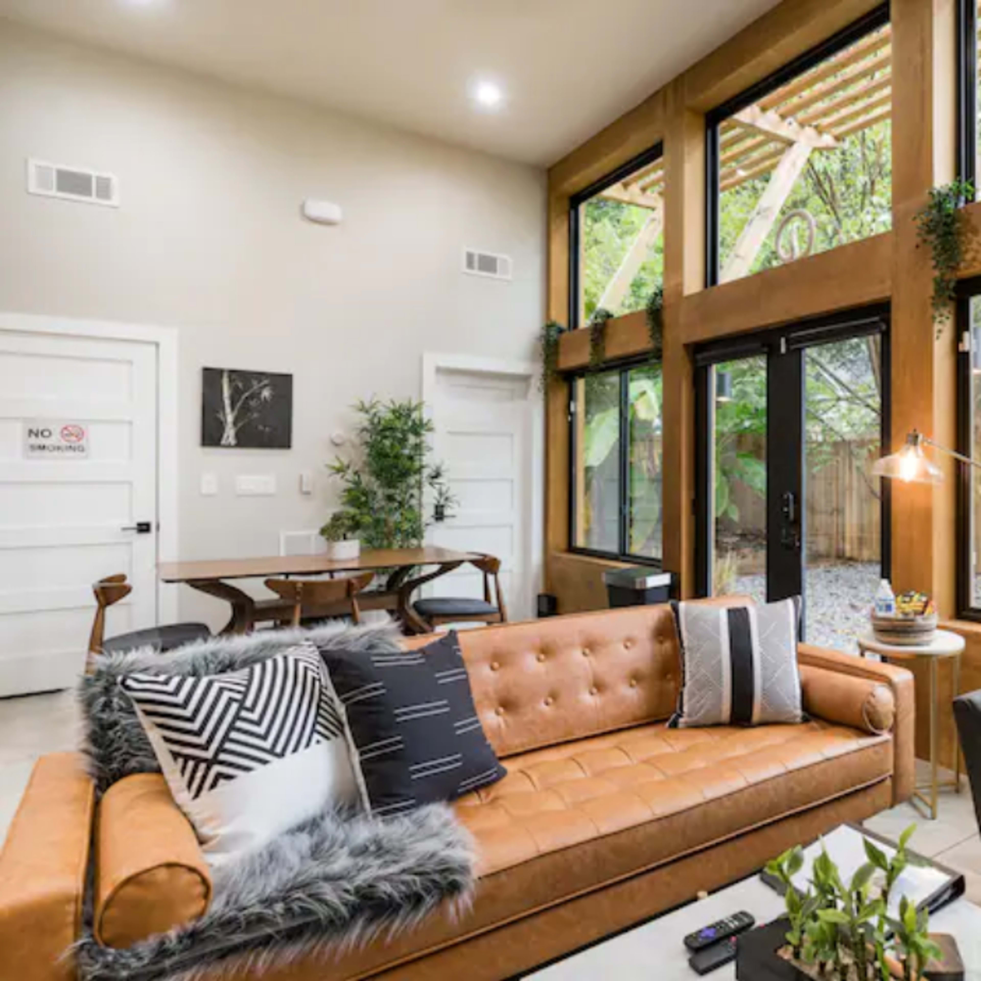 A modern living room features a leather sofa, a gray armchair, and large windows overlooking a patio and trees.