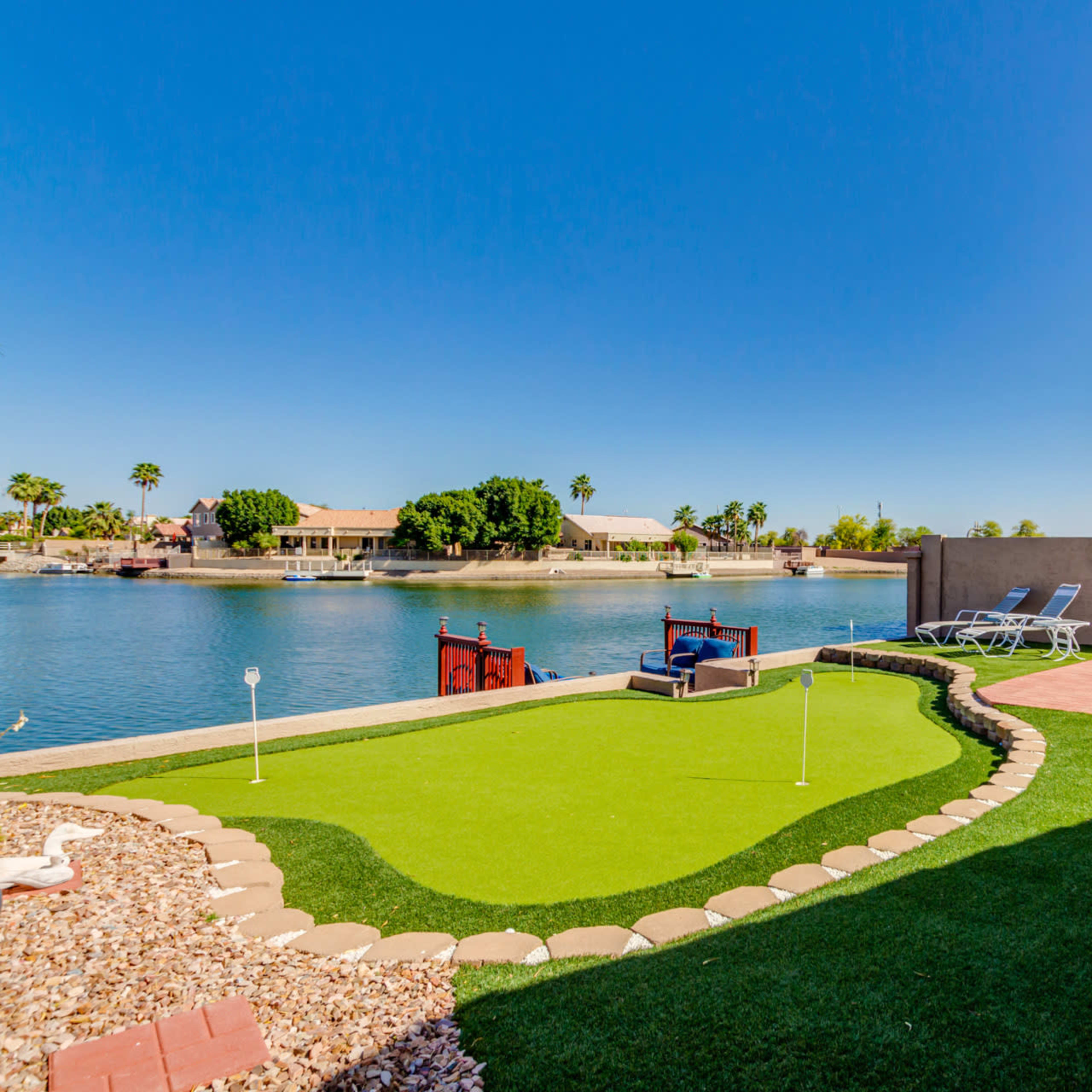 A backyard features a putting green, patio furniture, and a view of the water and houses across the canal.