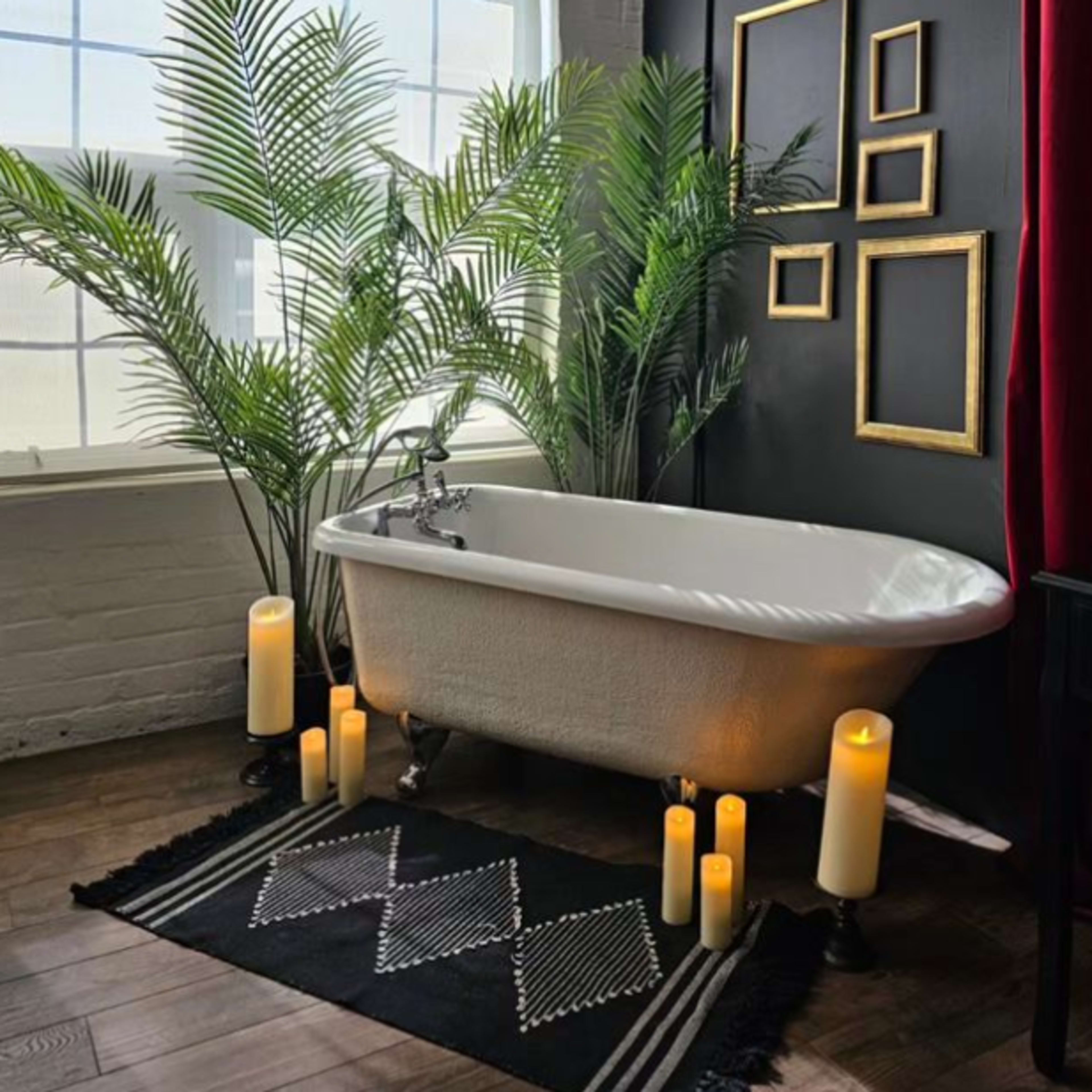 A clawfoot bathtub surrounded by tall plants and lit candles, set against a backdrop of a black wall and a window.