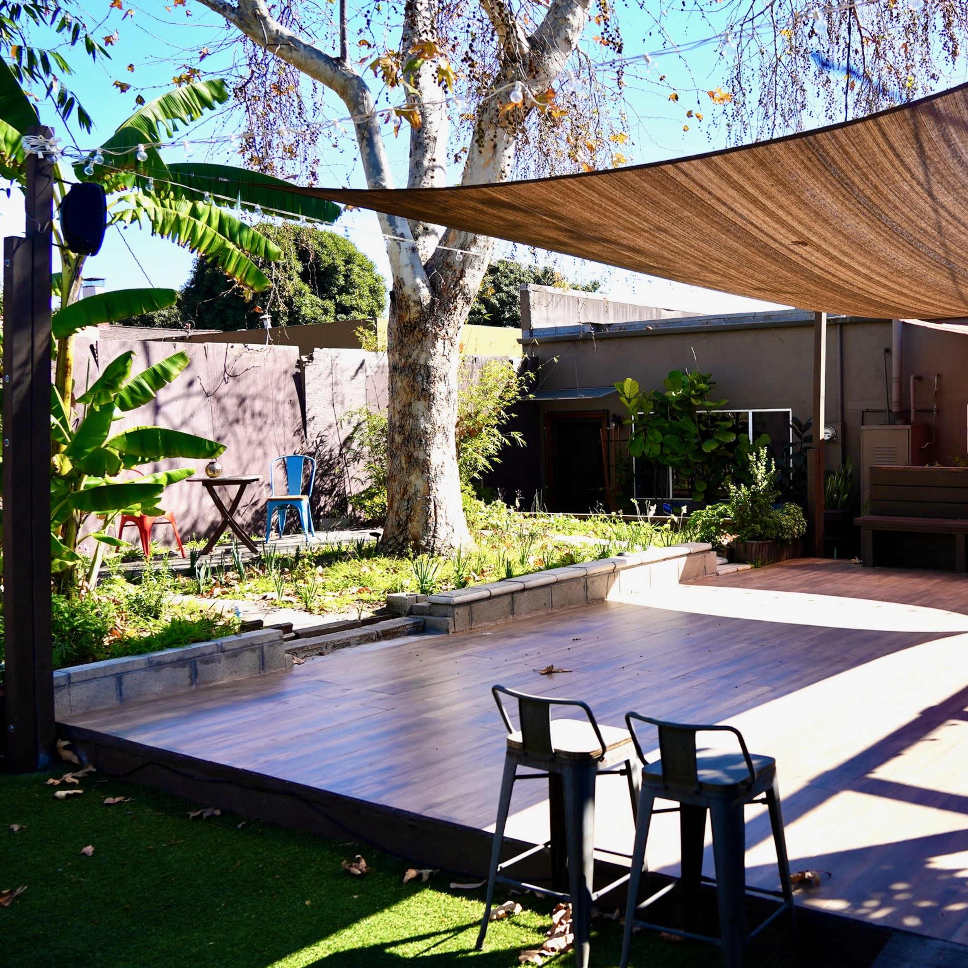 A wooden deck area shaded by a large fabric canopy, surrounded by greenery and buildings.