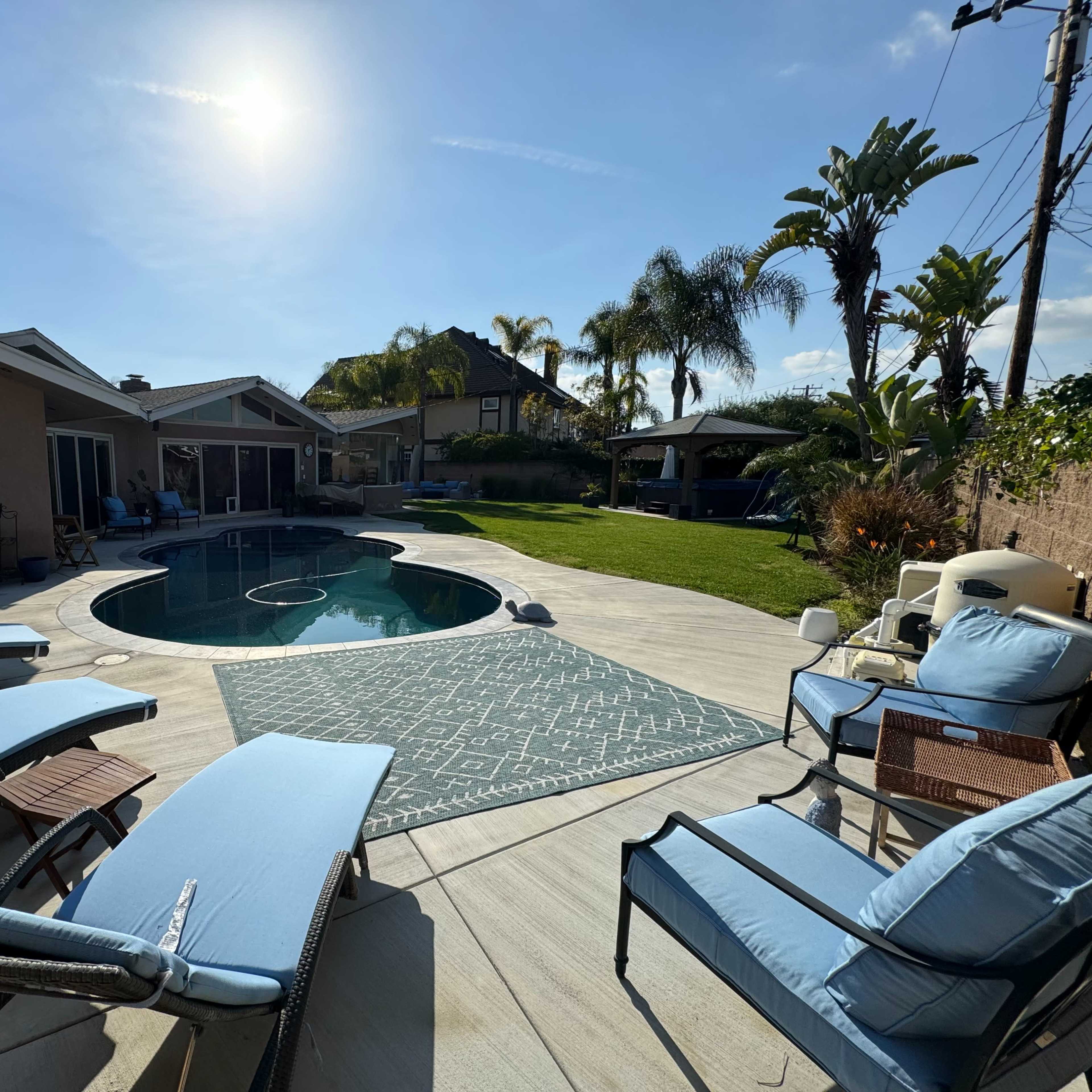 A pool area with lounge chairs, a circular pool, and surrounding landscaped lawn and palm trees under a clear sky.