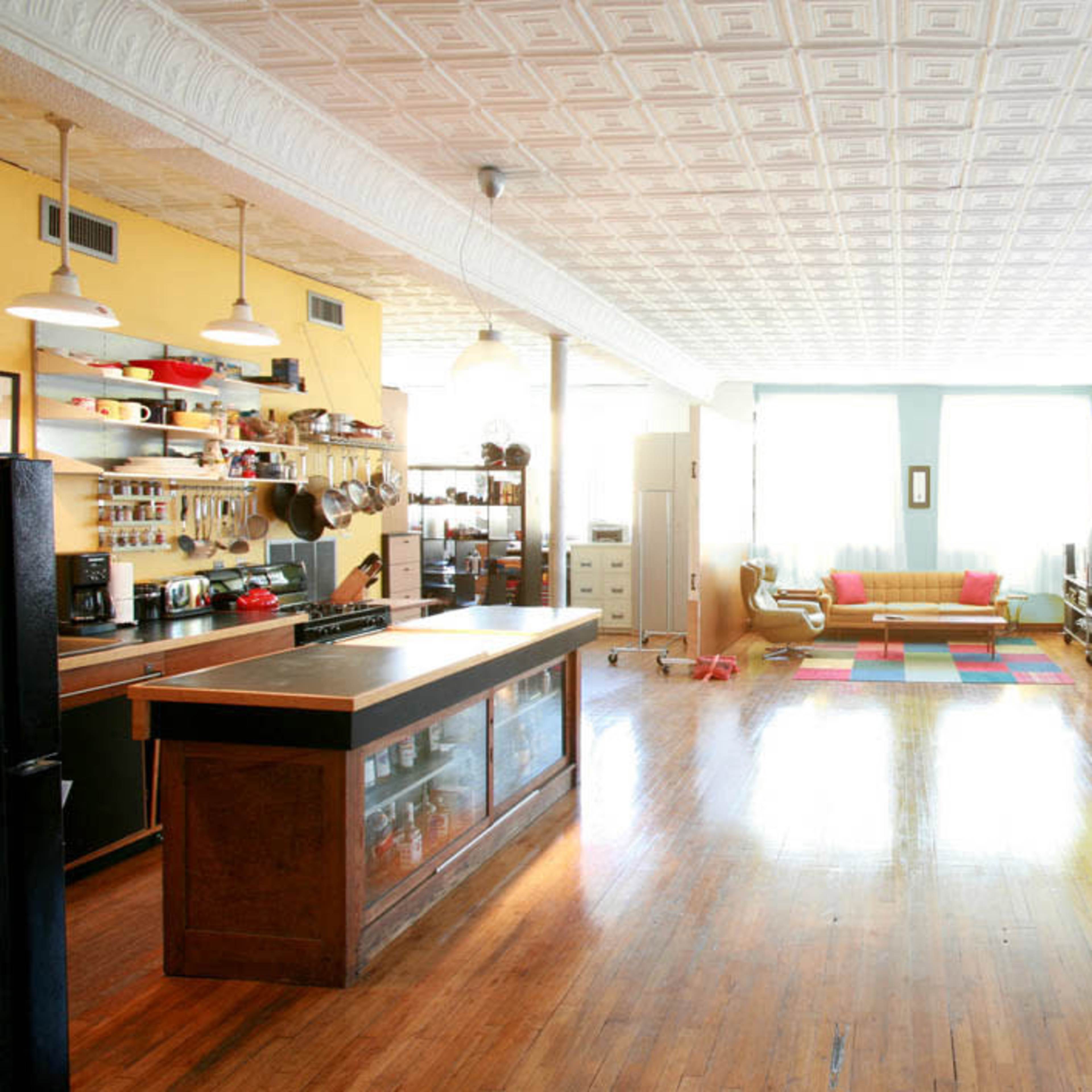 A spacious, bright room featuring a kitchen area with a counter, a cozy seating area with a sofa, and a colorful rug on the wooden floor.