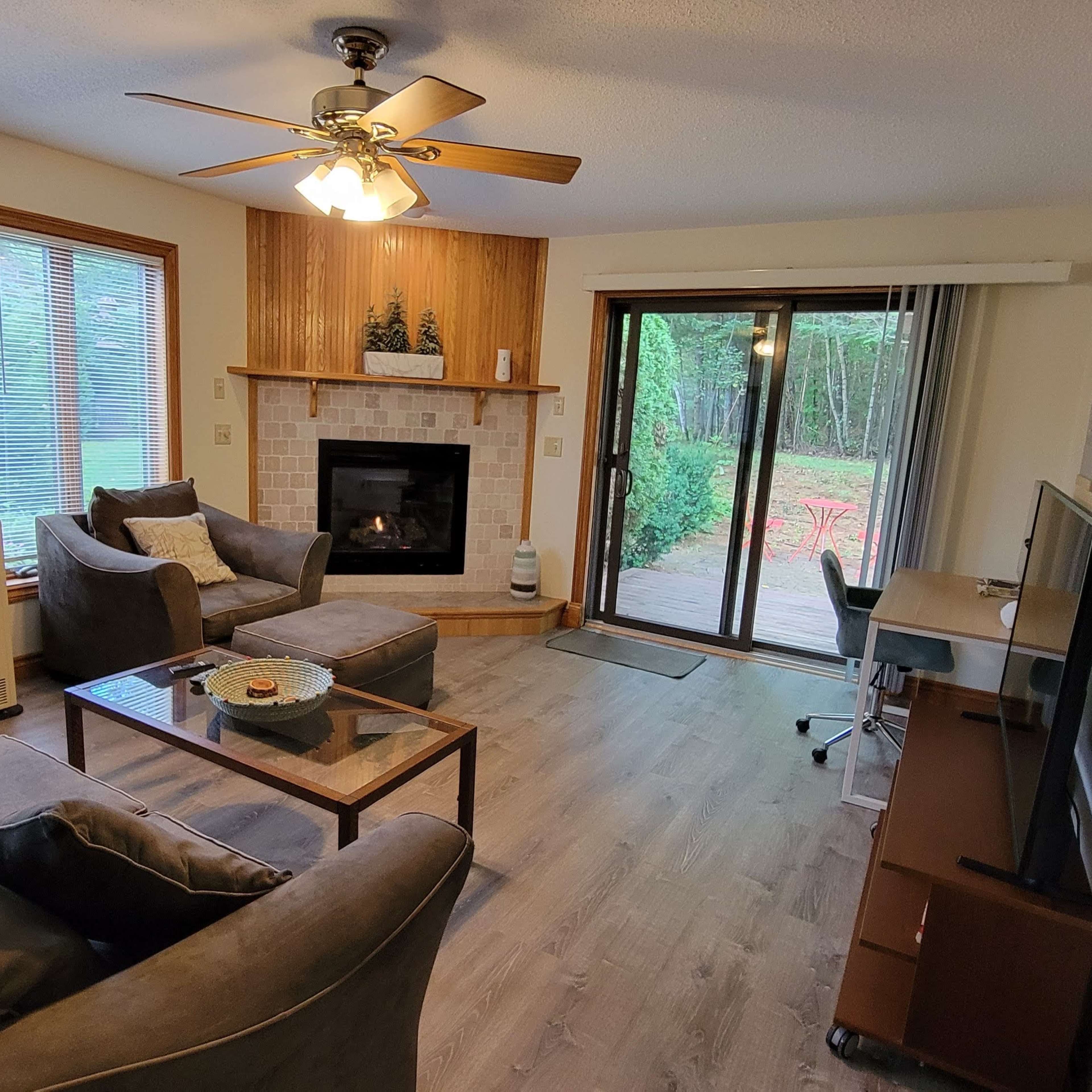 A cozy living room with a sectional sofa, a coffee table, a television, and a sliding glass door leading to a patio surrounded by trees.