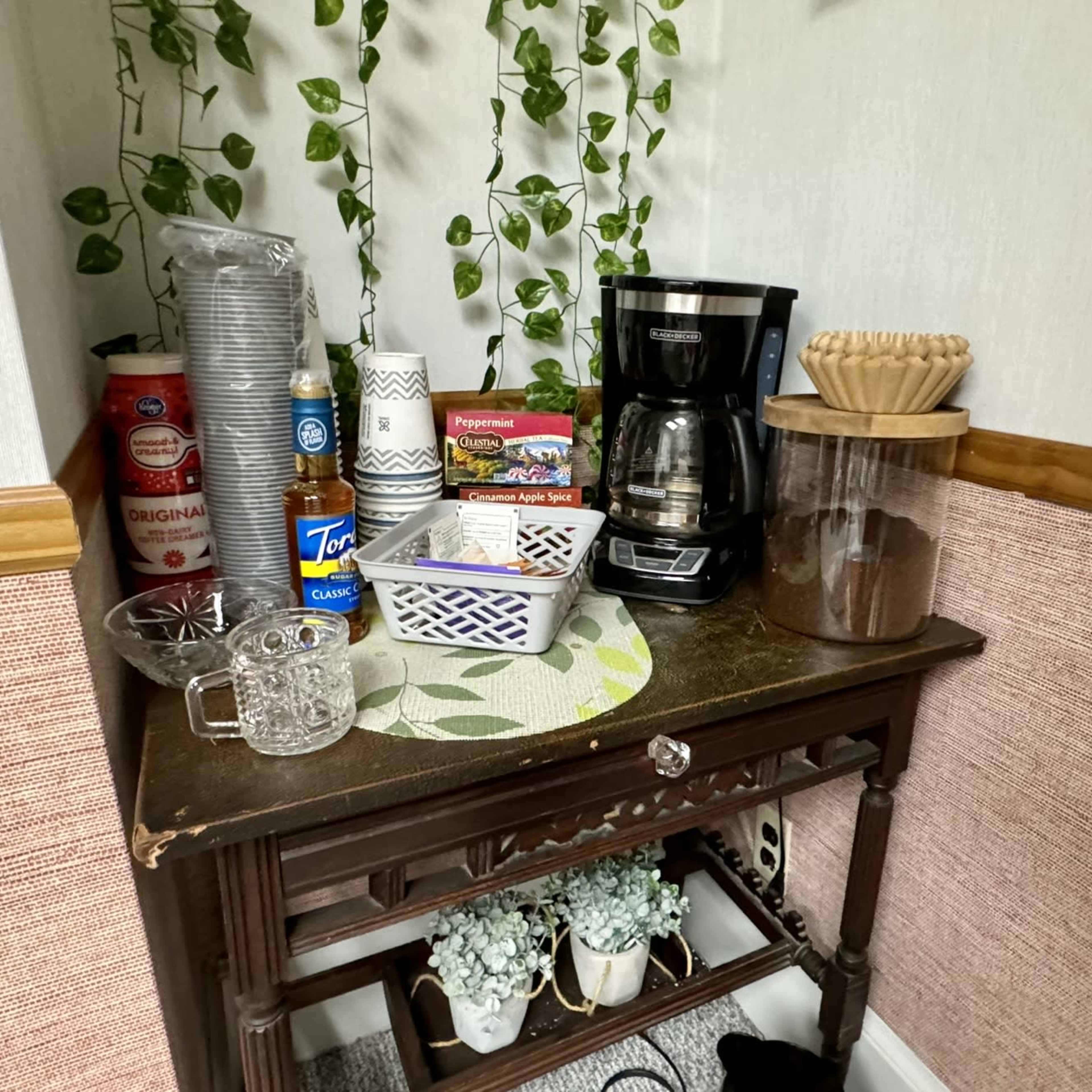 A corner table with a coffee maker, several containers, and glasses, surrounded by green plants on the wall.
