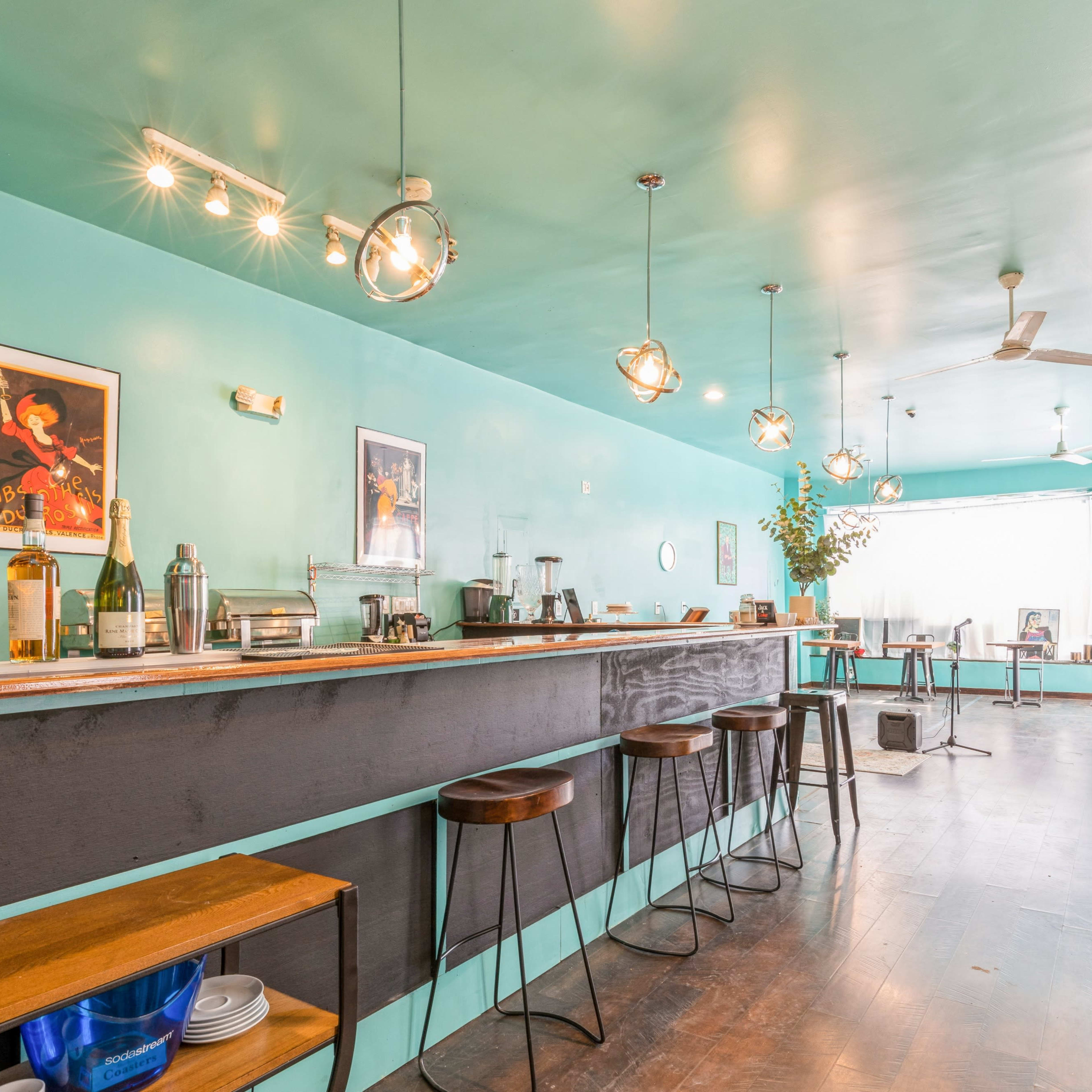 A modern bar with a turquoise wall, wooden stools, and pendant lights, featuring a counter with various bar tools and bottles.