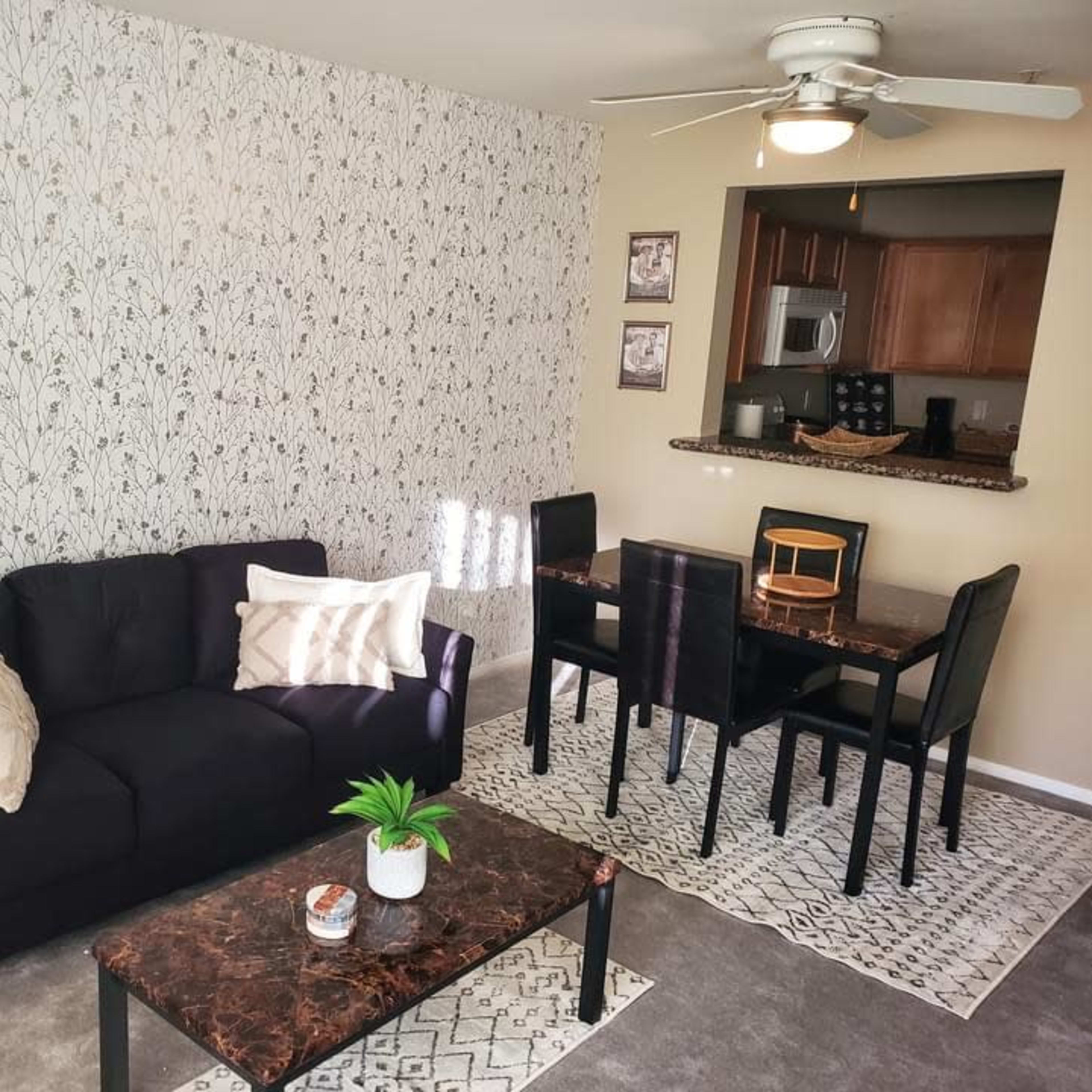 A living room with a black sofa, patterned coffee table, and a dining area in the background featuring a round wooden table and black chairs.