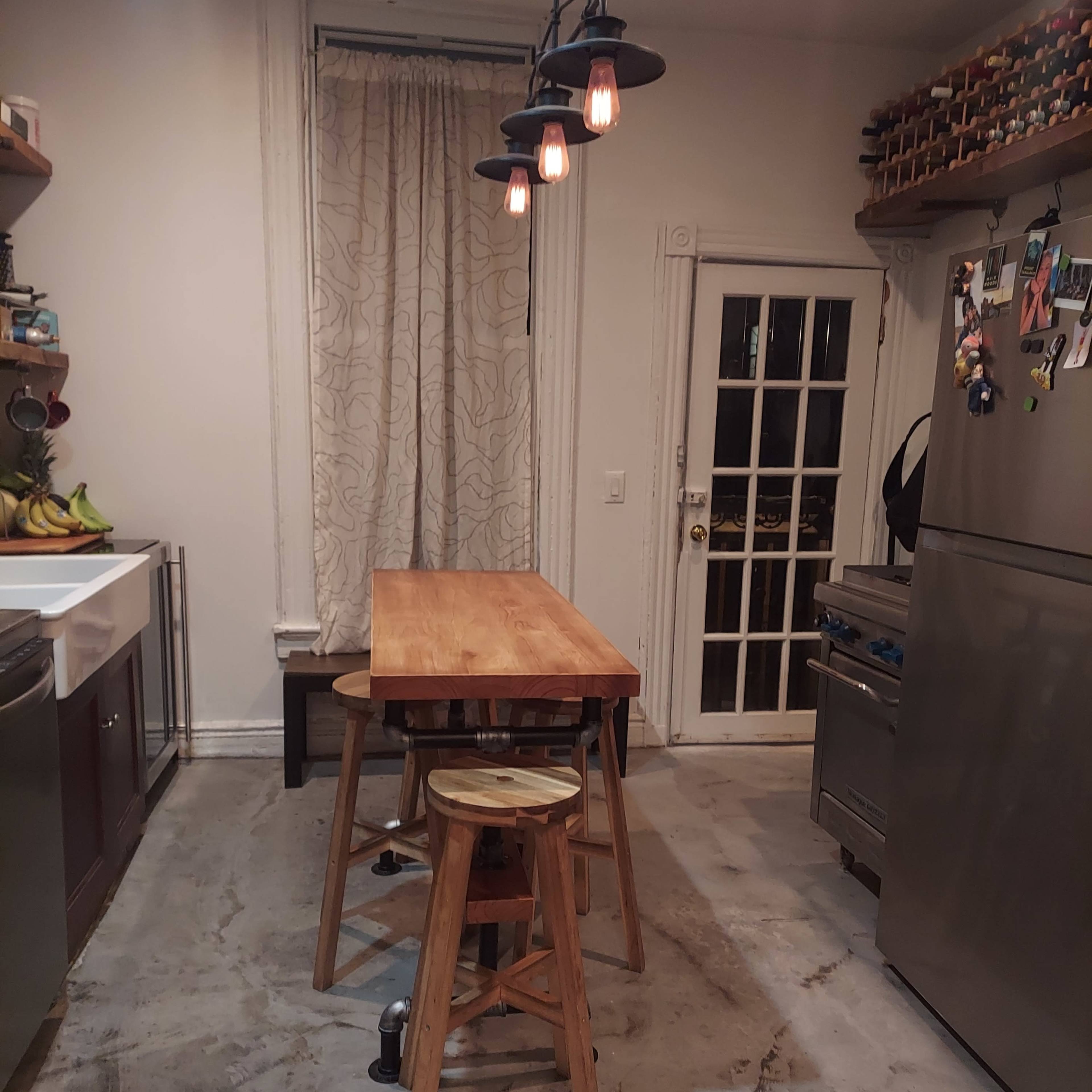 A wooden table with stools is positioned in a kitchen featuring a gray tiled floor and a single light fixture hanging above.