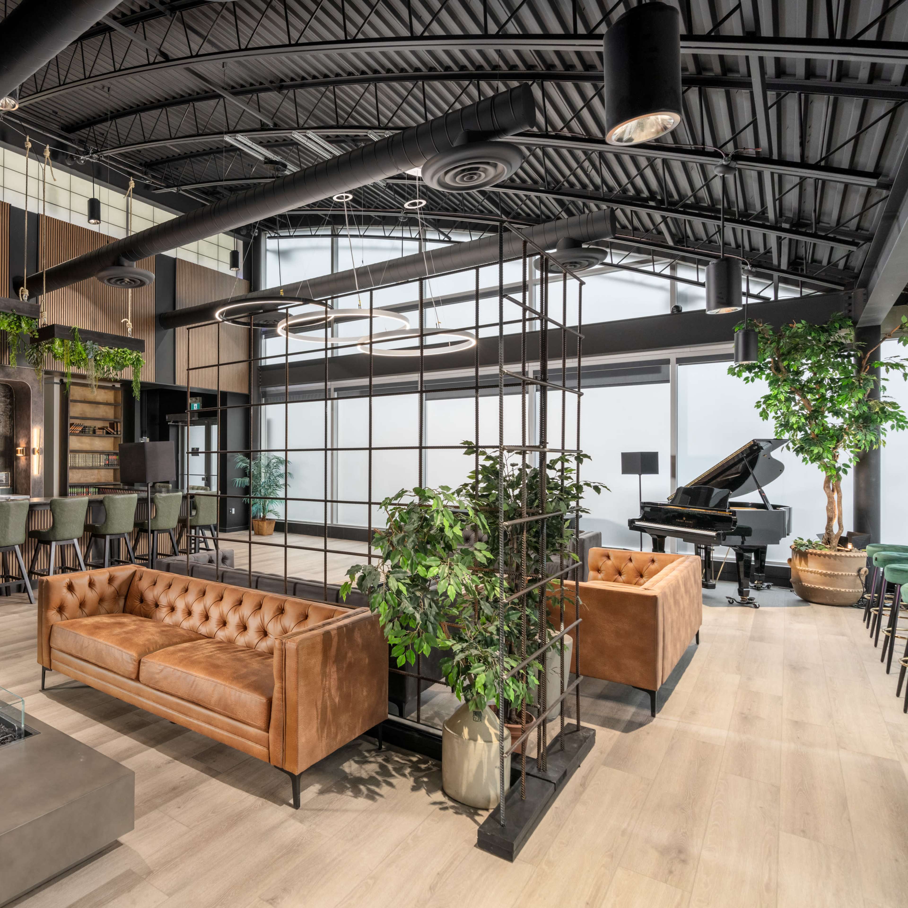 A modern lounge area featuring a bar with seating, a piano in the corner, and brown leather sofas arranged around a central fire pit.