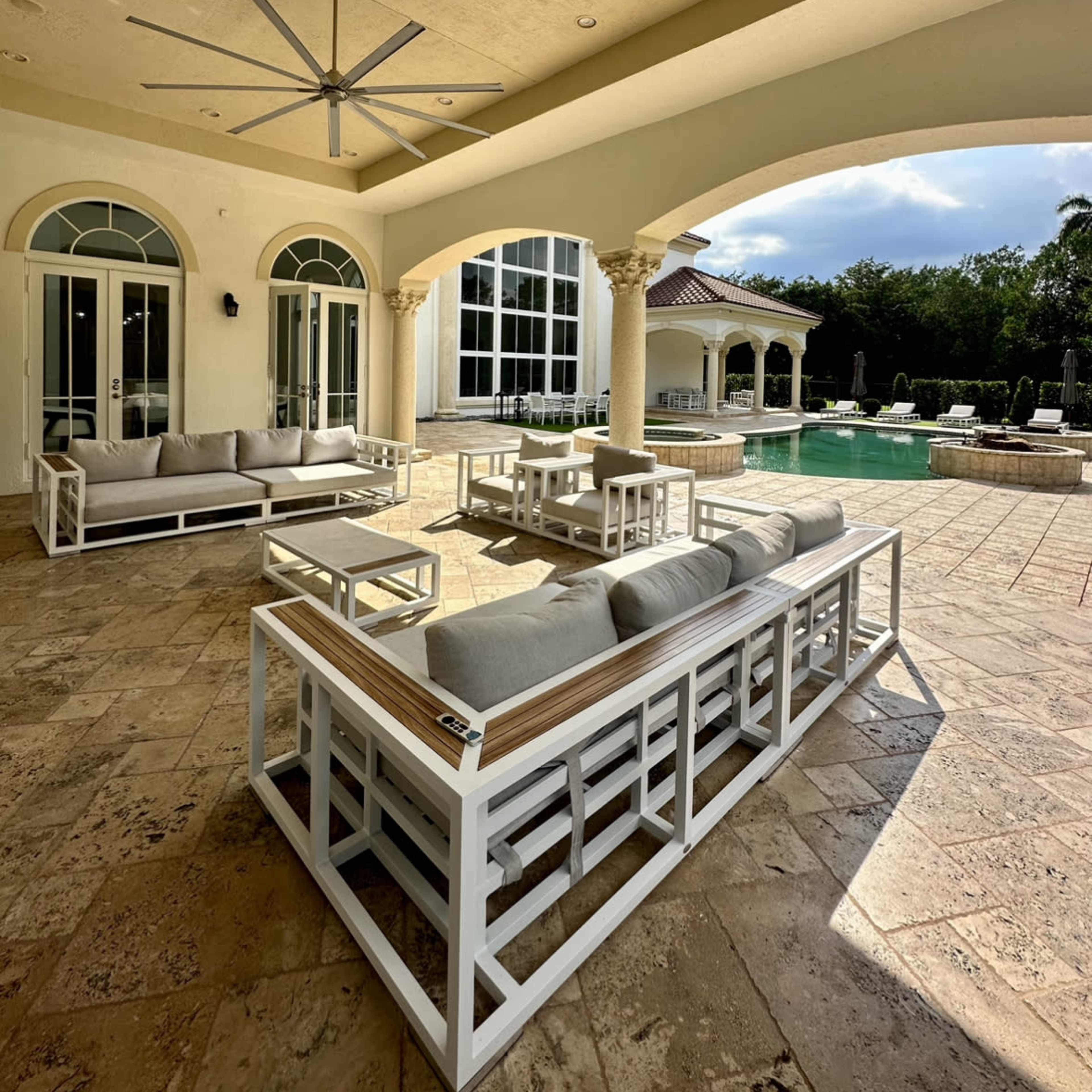 A patio features white outdoor furniture arranged near a swimming pool, with large windows and columns in the background.