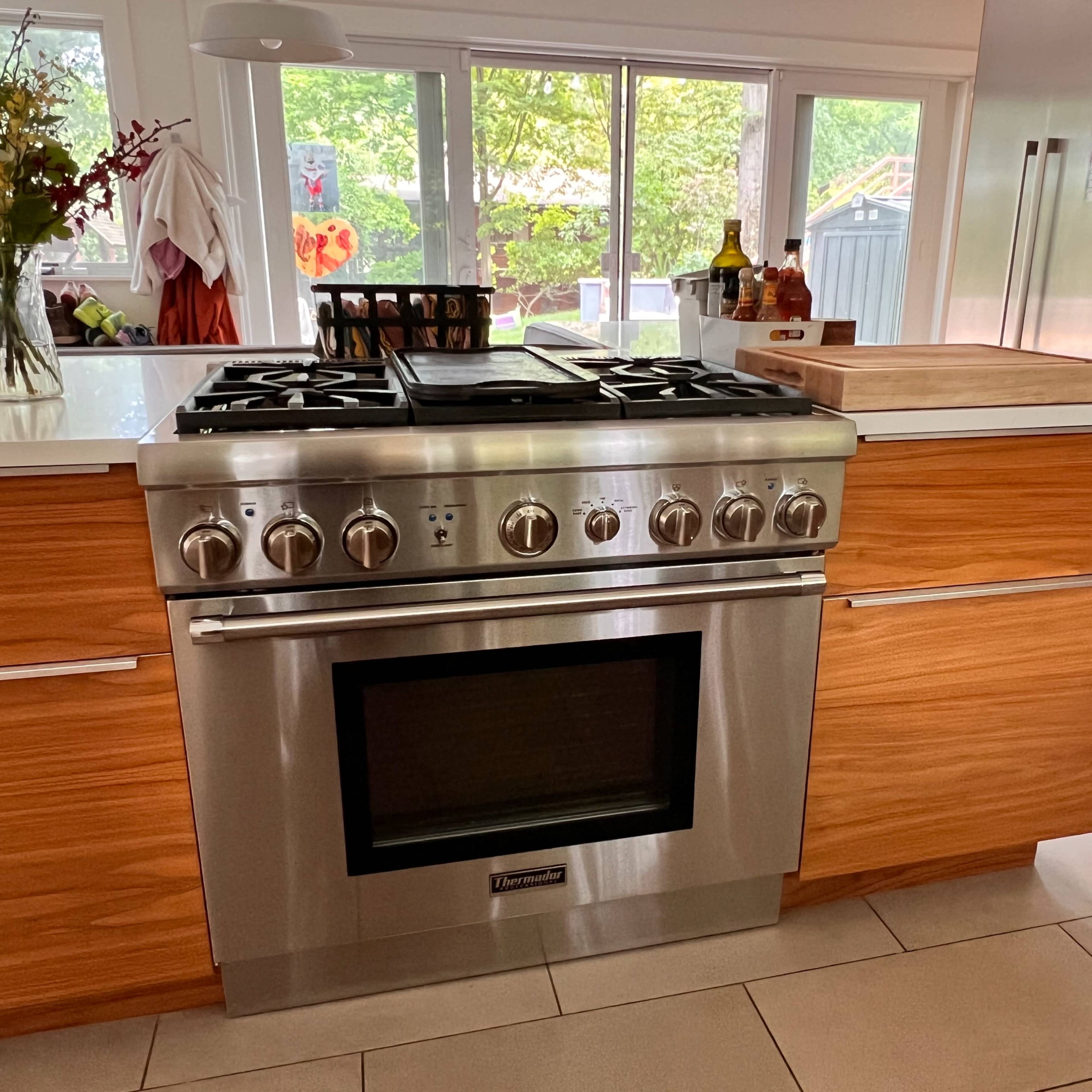 A modern kitchen featuring a stainless steel gas range with six burners, positioned between wooden cabinetry and illuminated by natural light from large windows.